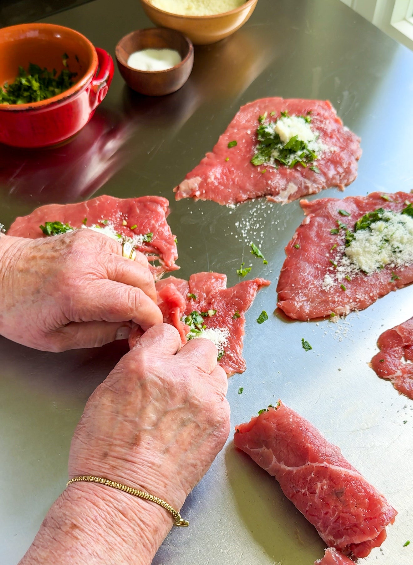 Hands rolling thin slices of raw beef stuffed with chopped herbs, grated cheese, and seasoning on a metal surface, with bowls of parsley and salt in the background.