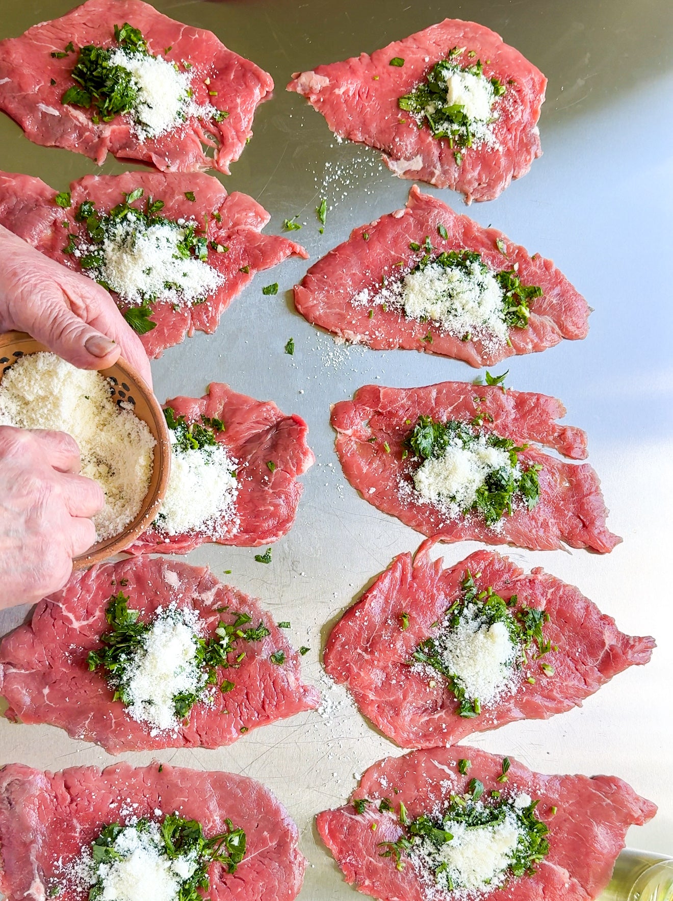 Slices of raw beef laid out on a metal surface, each topped with chopped herbs and grated cheese. A persons hands are sprinkling more cheese onto one of the slices from a small bowl.