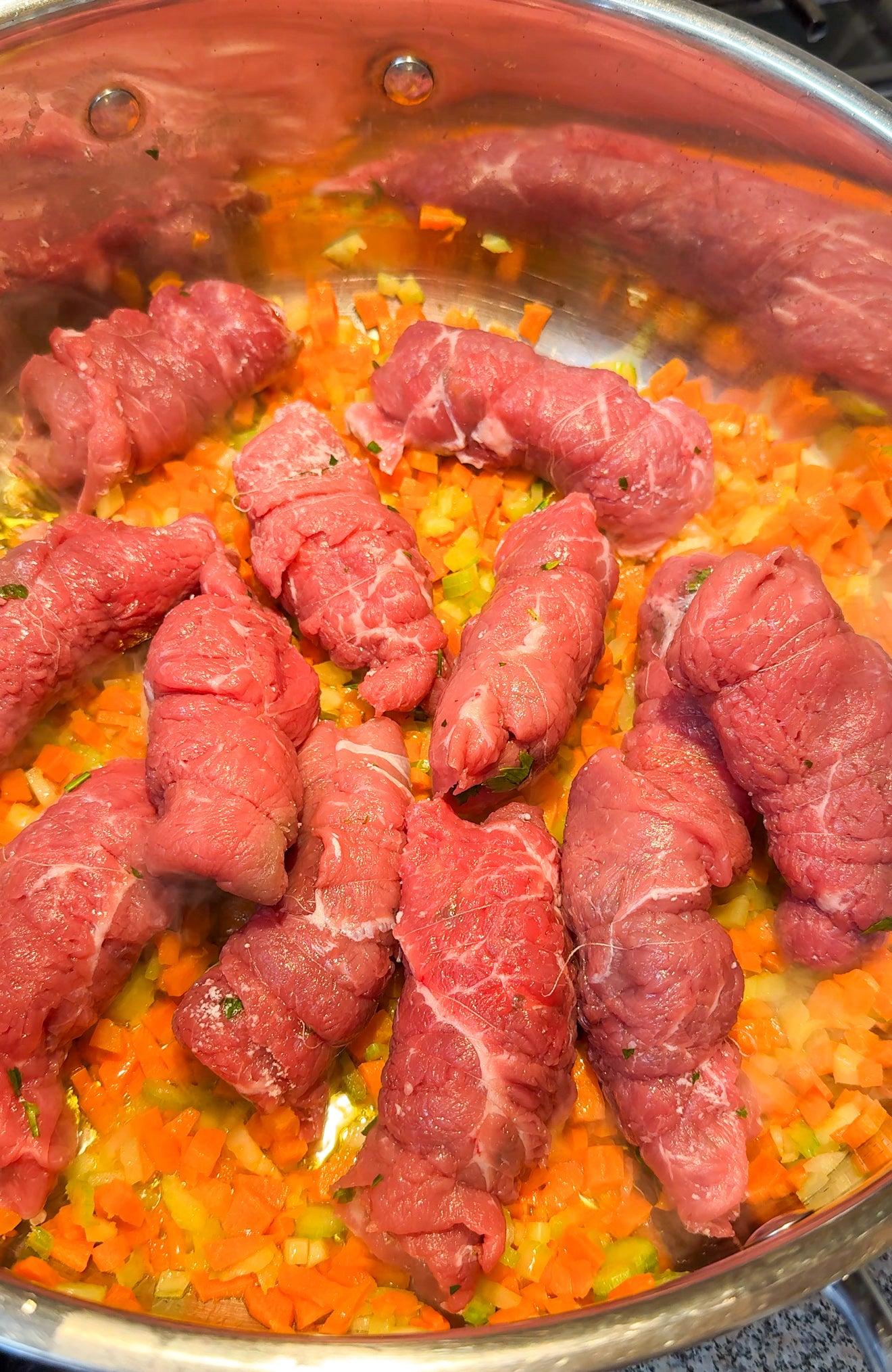 Raw beef rolls arranged in a large stainless steel pan on top of diced carrots and celery, ready to be cooked.