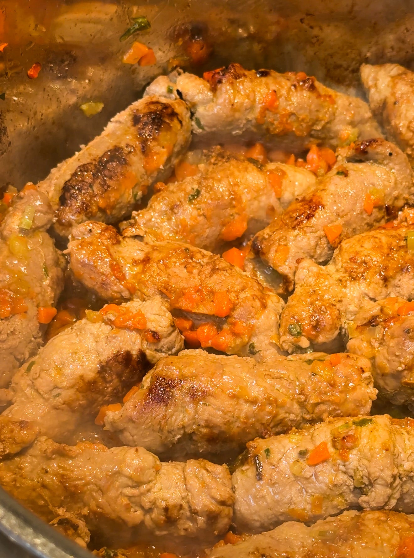 A close-up of browned meat rolls simmering in a pot with chopped carrots, onions, and herbs, creating a hearty and flavorful dish. Steam rises from the mixture.