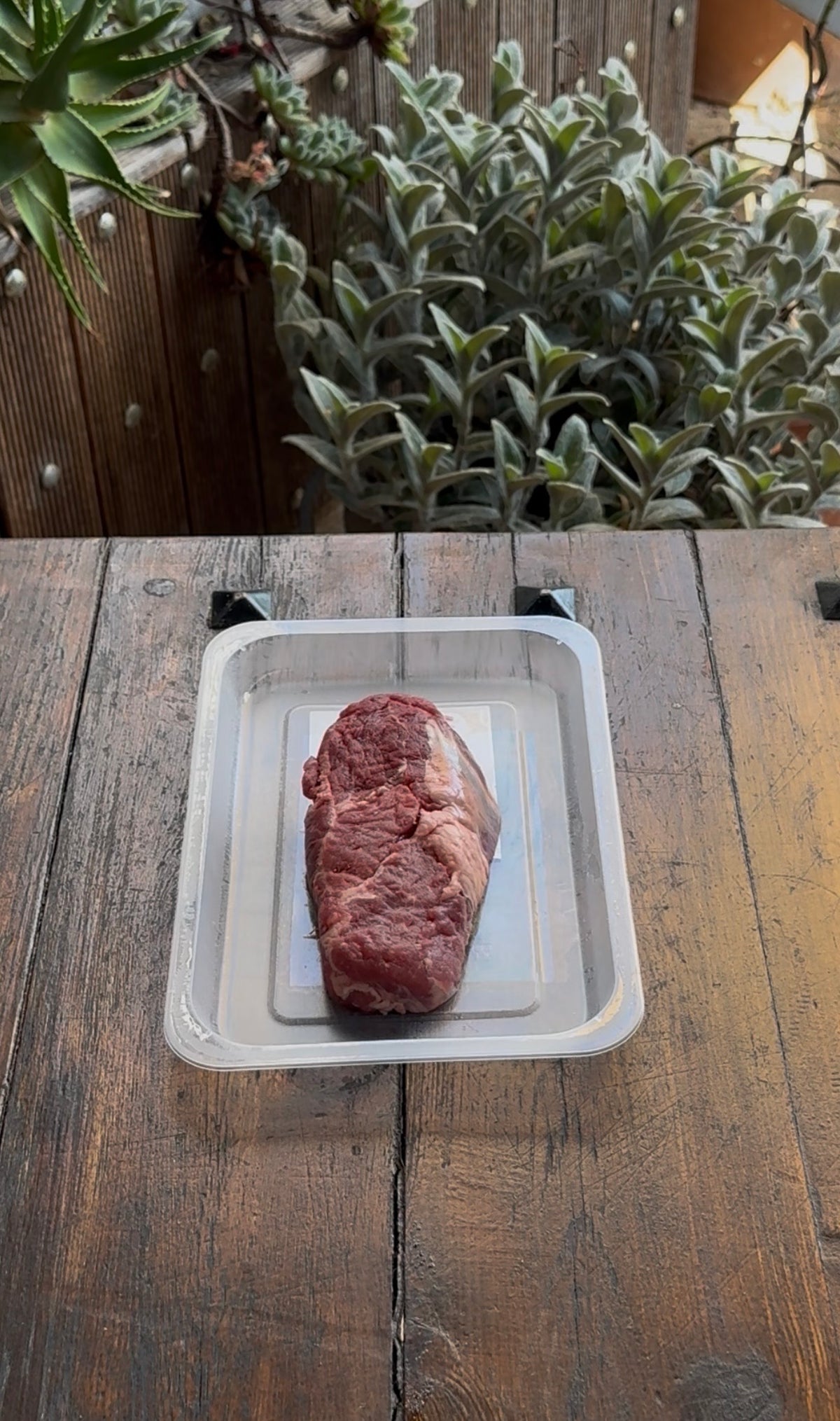 A raw steak in a plastic tray resting on a wooden table outdoors.