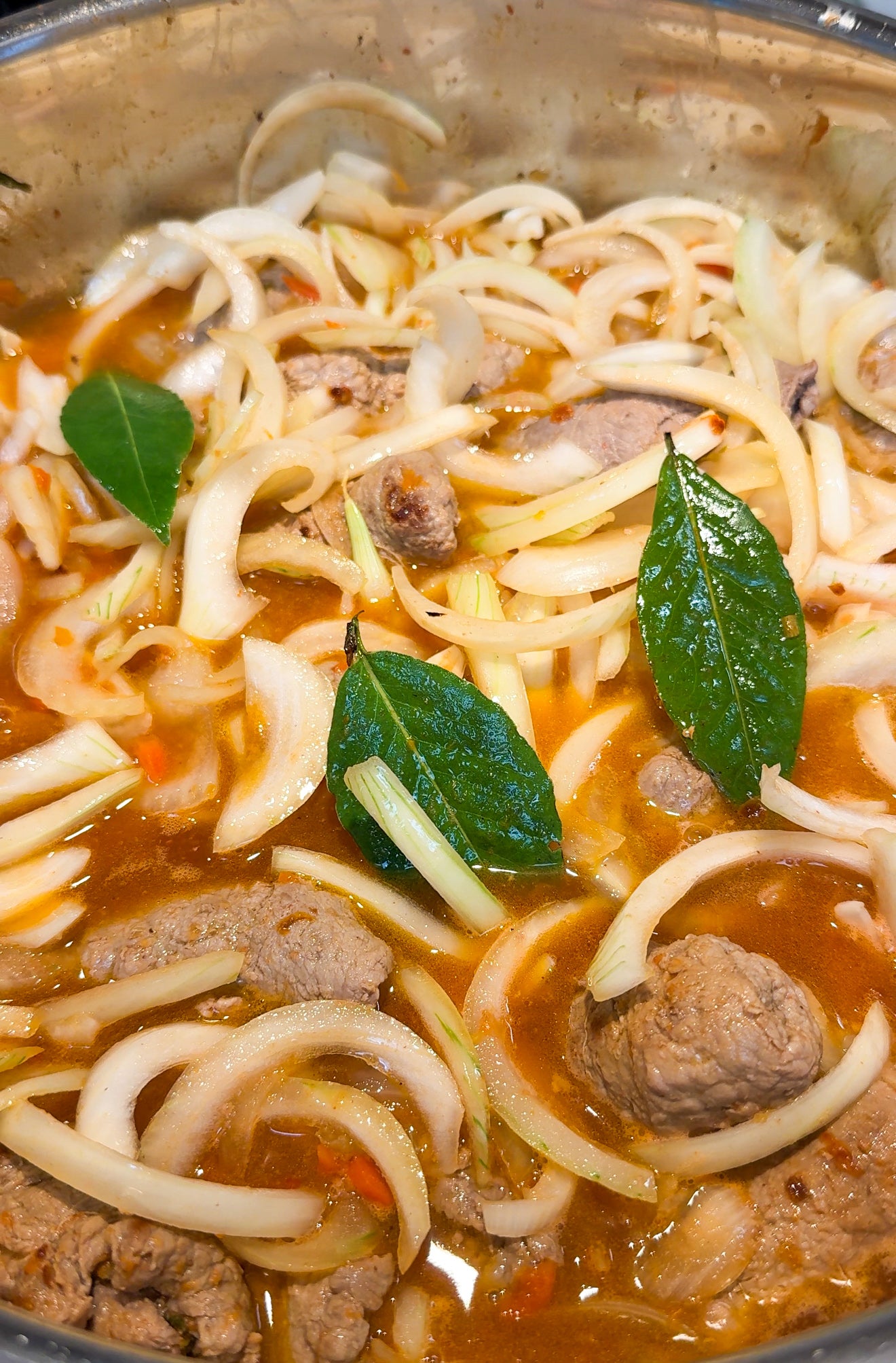 A close-up of a pot filled with a stew containing beef chunks, sliced onions, bay leaves, and a rich, reddish broth.