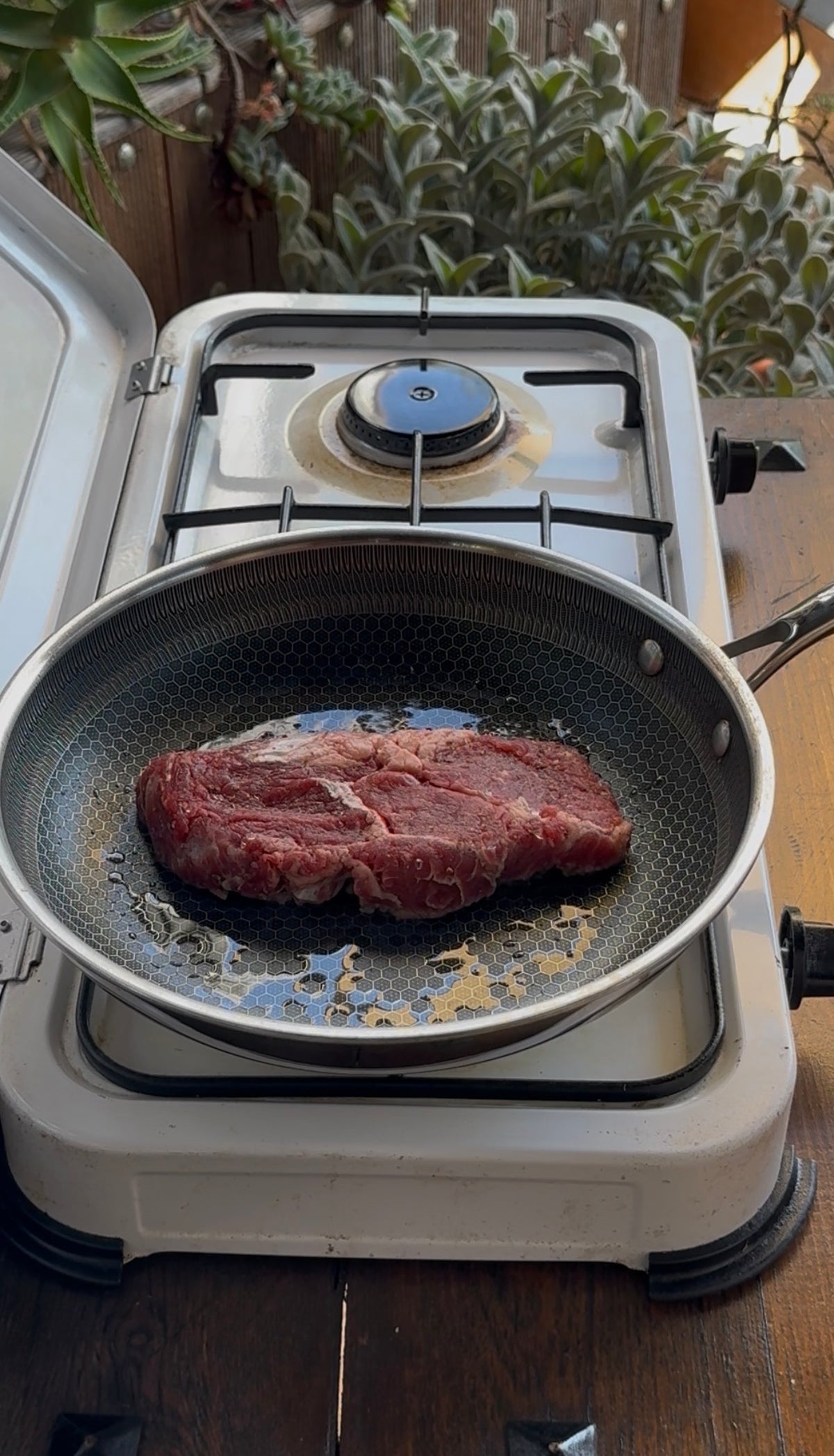 A raw steak cooking in a non-stick frying pan on a portable gas stove.