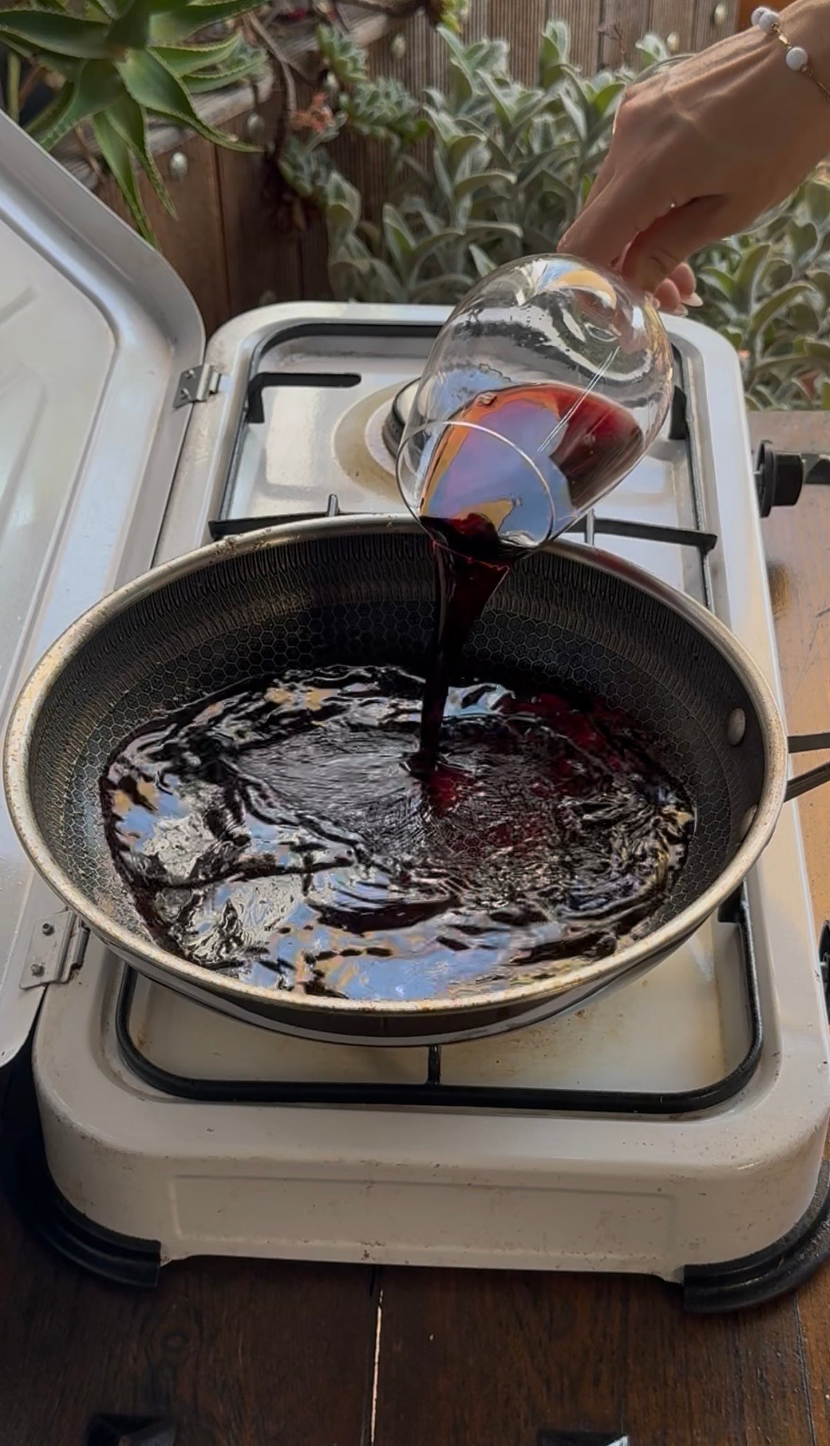 A hand pours red wine from a glass into a wide skillet on a stovetop.