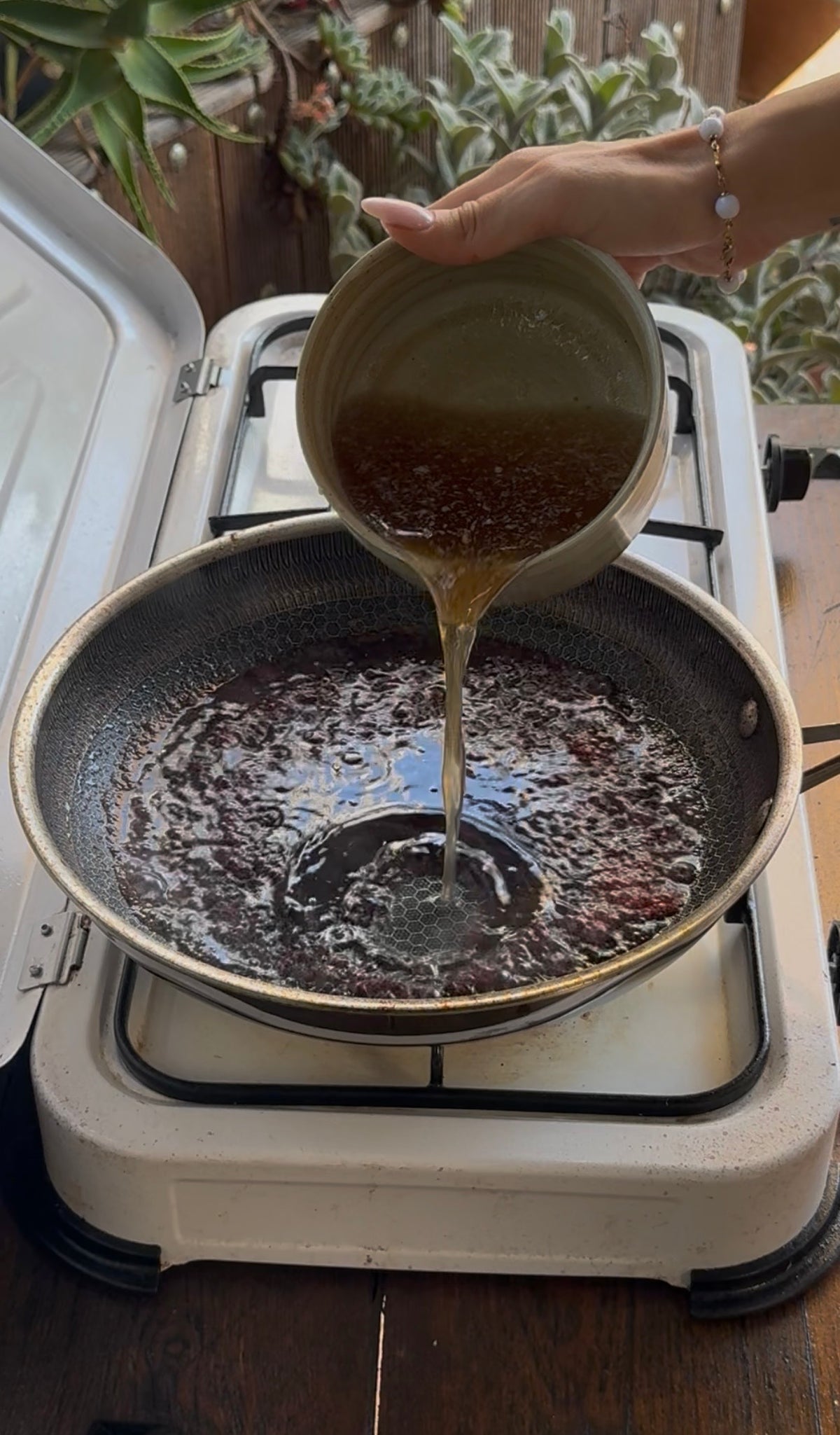 A hand pours liquid from a small bowl into a frying pan containing dark, simmering fruit.