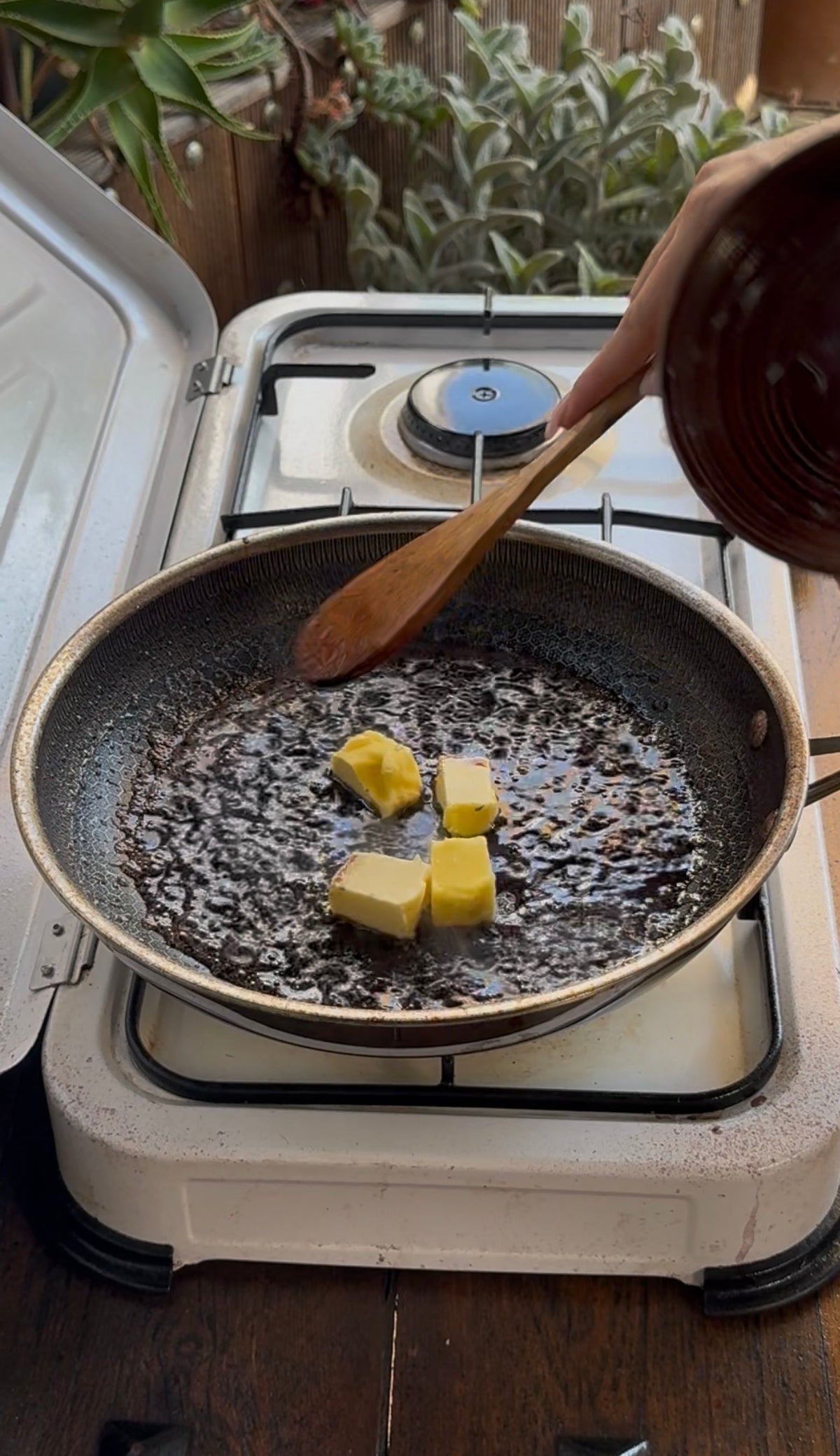 A person uses a wooden spoon to stir four cubes of butter melting in a hot frying pan on a stovetop.