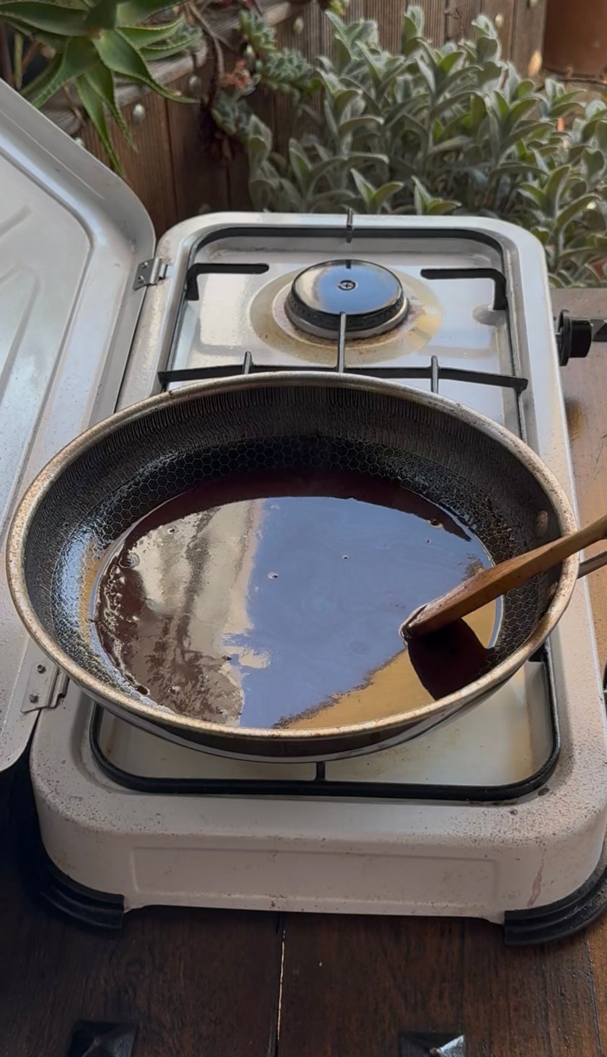 A dark liquid simmering in a frying pan on a portable stove, with a wooden spoon resting inside.