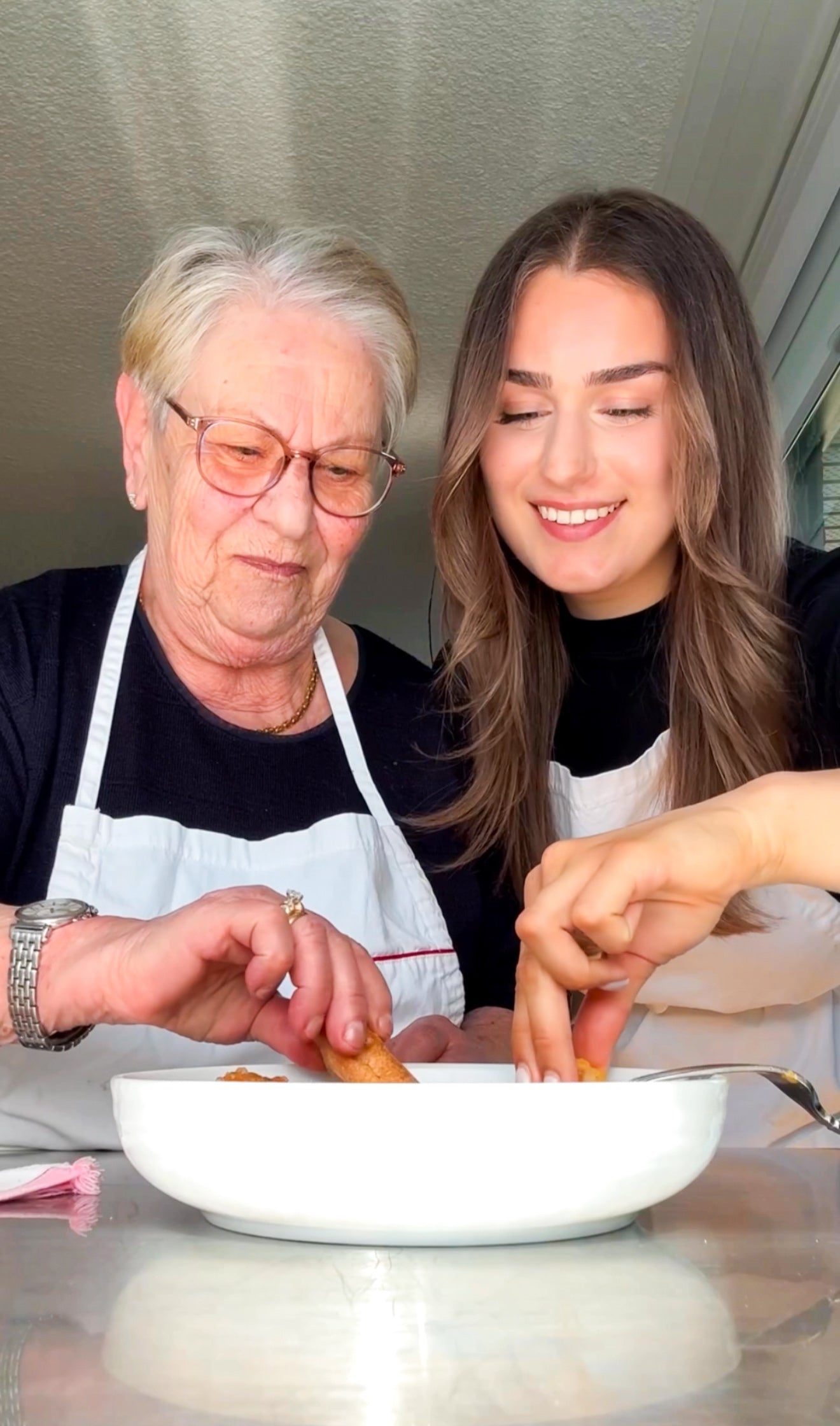 An elderly woman and a young woman, both wearing aprons, smile as they prepare food together, dipping pieces into a white dish. The scene suggests a warm, shared cooking moment in a bright kitchen.