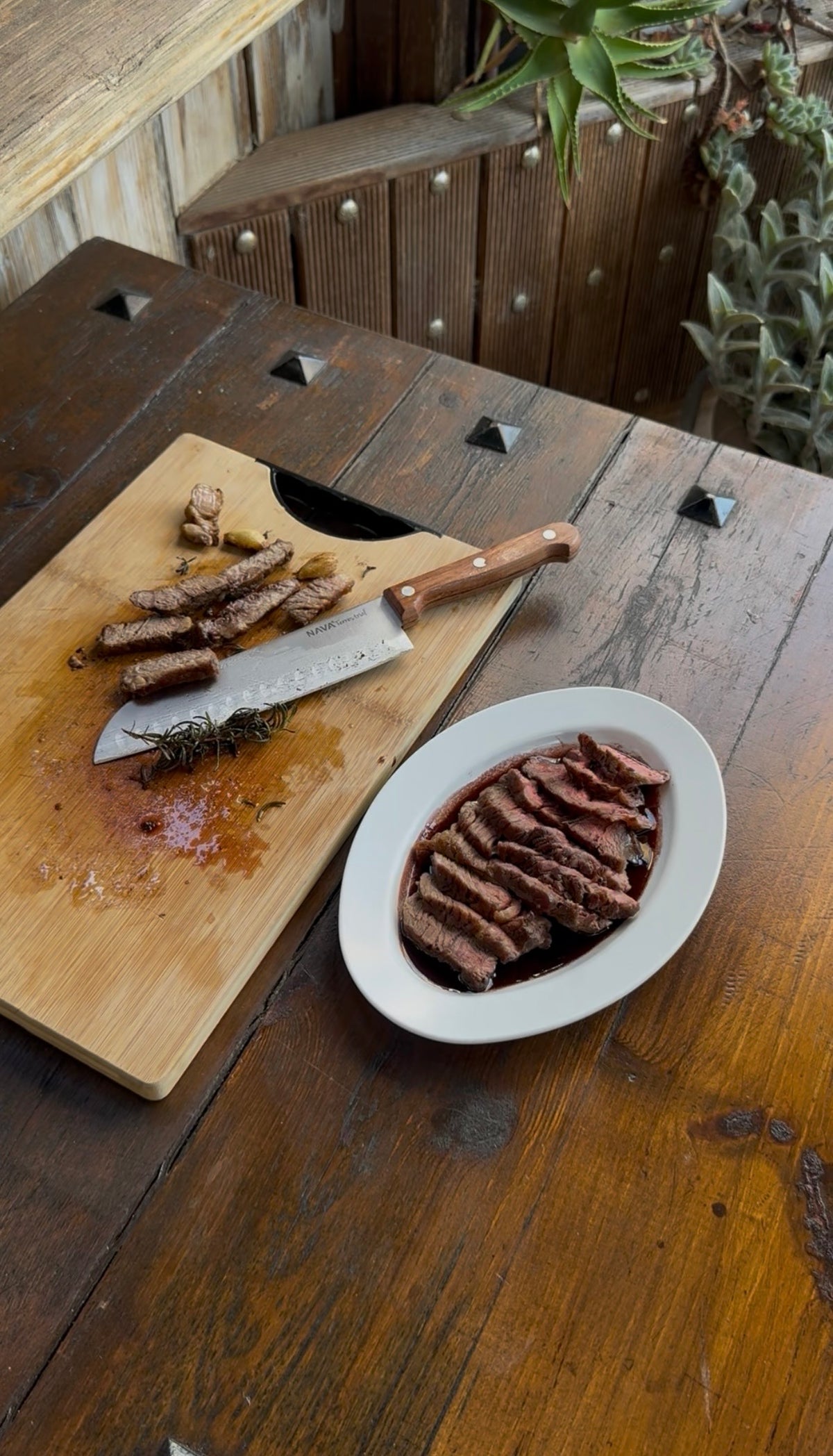 A sliced steak served on a white platter next to a wooden cutting board with a chefs knife, rosemary, and garlic cloves.