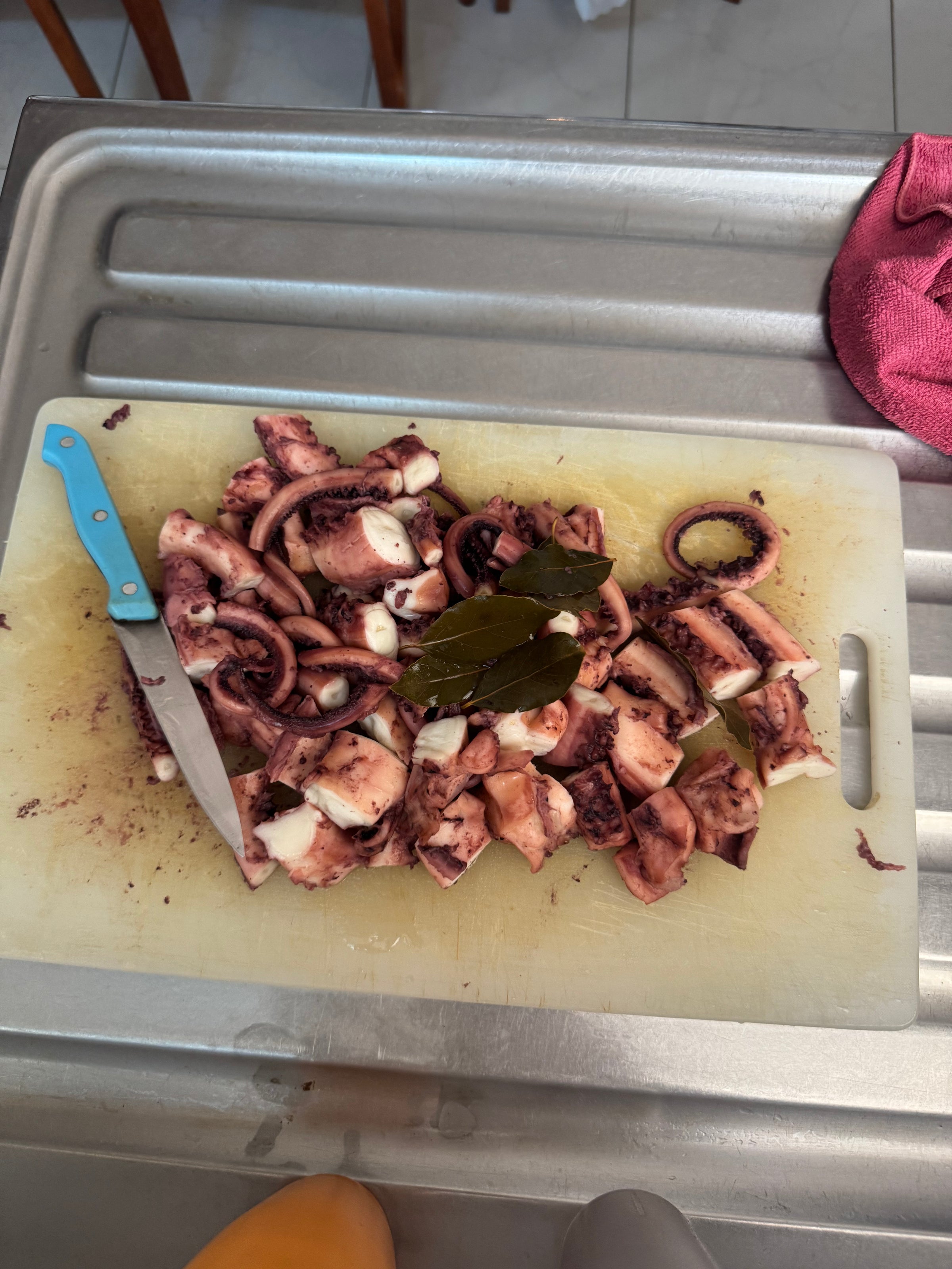 Chopped pieces of octopus and a few bay leaves on a cutting board with a knife, placed on a kitchen sink. A pink cloth is on the right side of the sink.