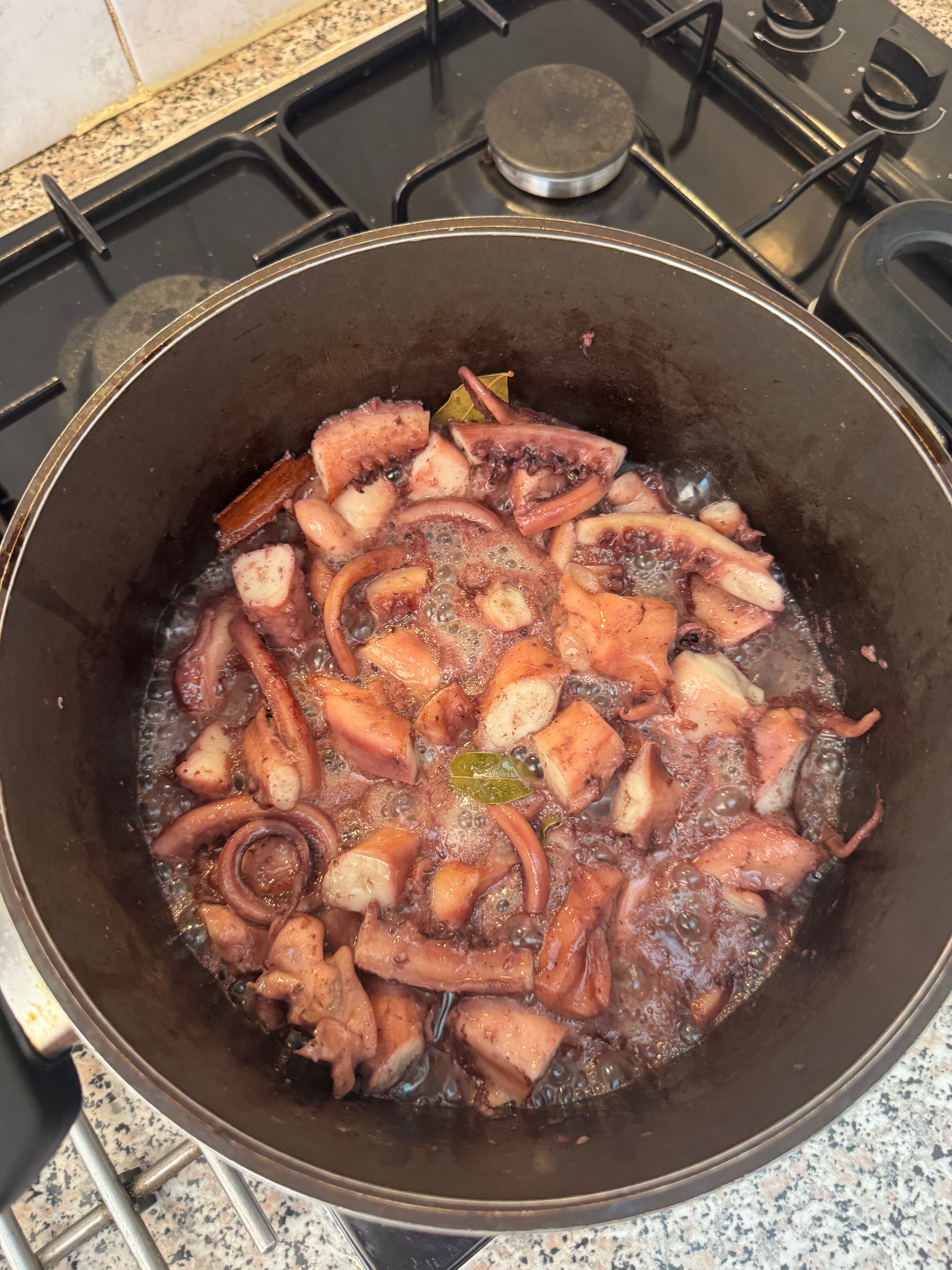 A pot on a stove contains chopped octopus simmering in a bubbling liquid with herbs and spices visible, such as bay leaf and cinnamon stick.
