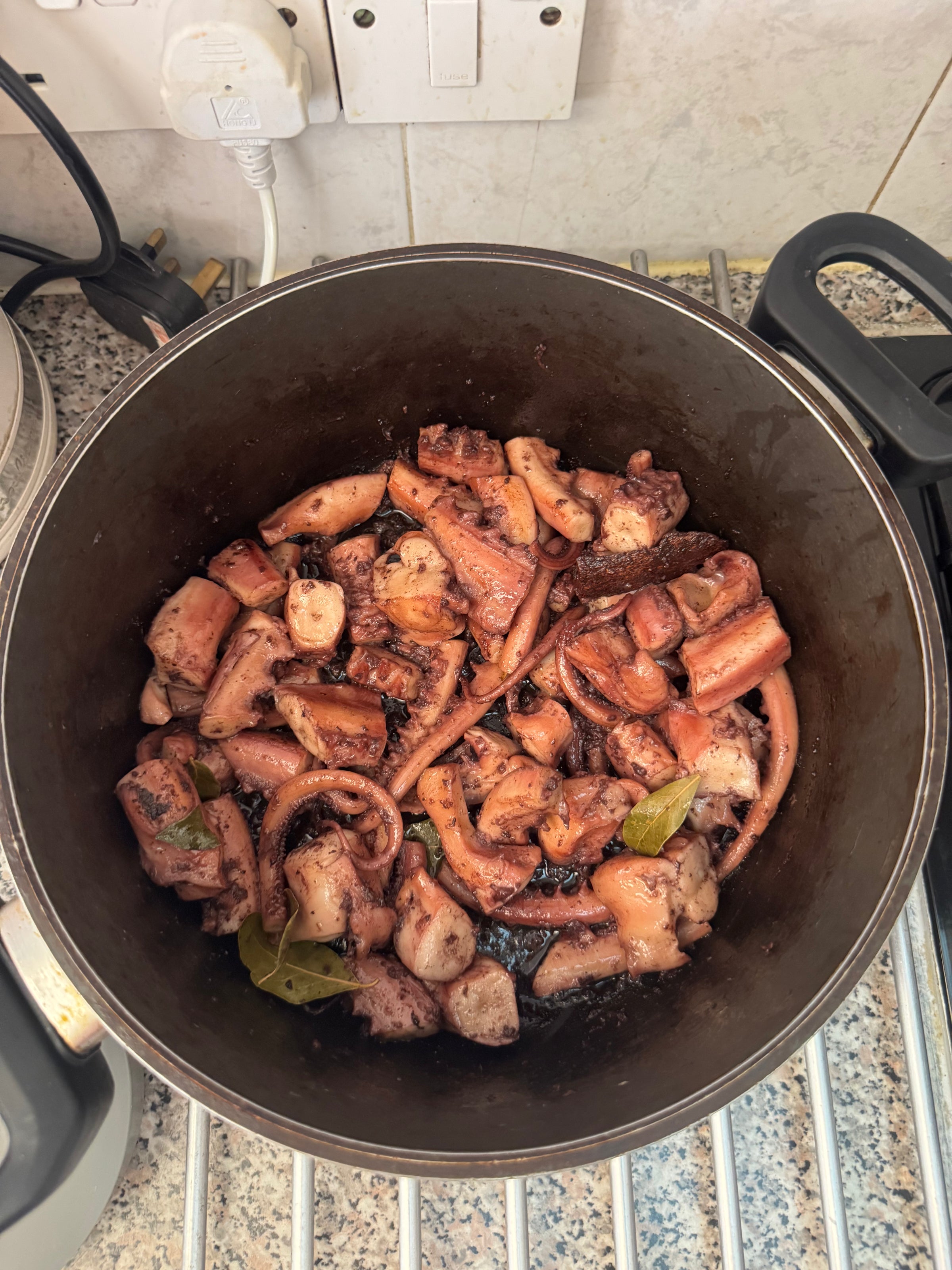 A pot filled with cooked pieces of octopus, some with visible tentacles, mixed with spices and bay leaves, sits on a kitchen stove with tiled walls and various appliances in the background.