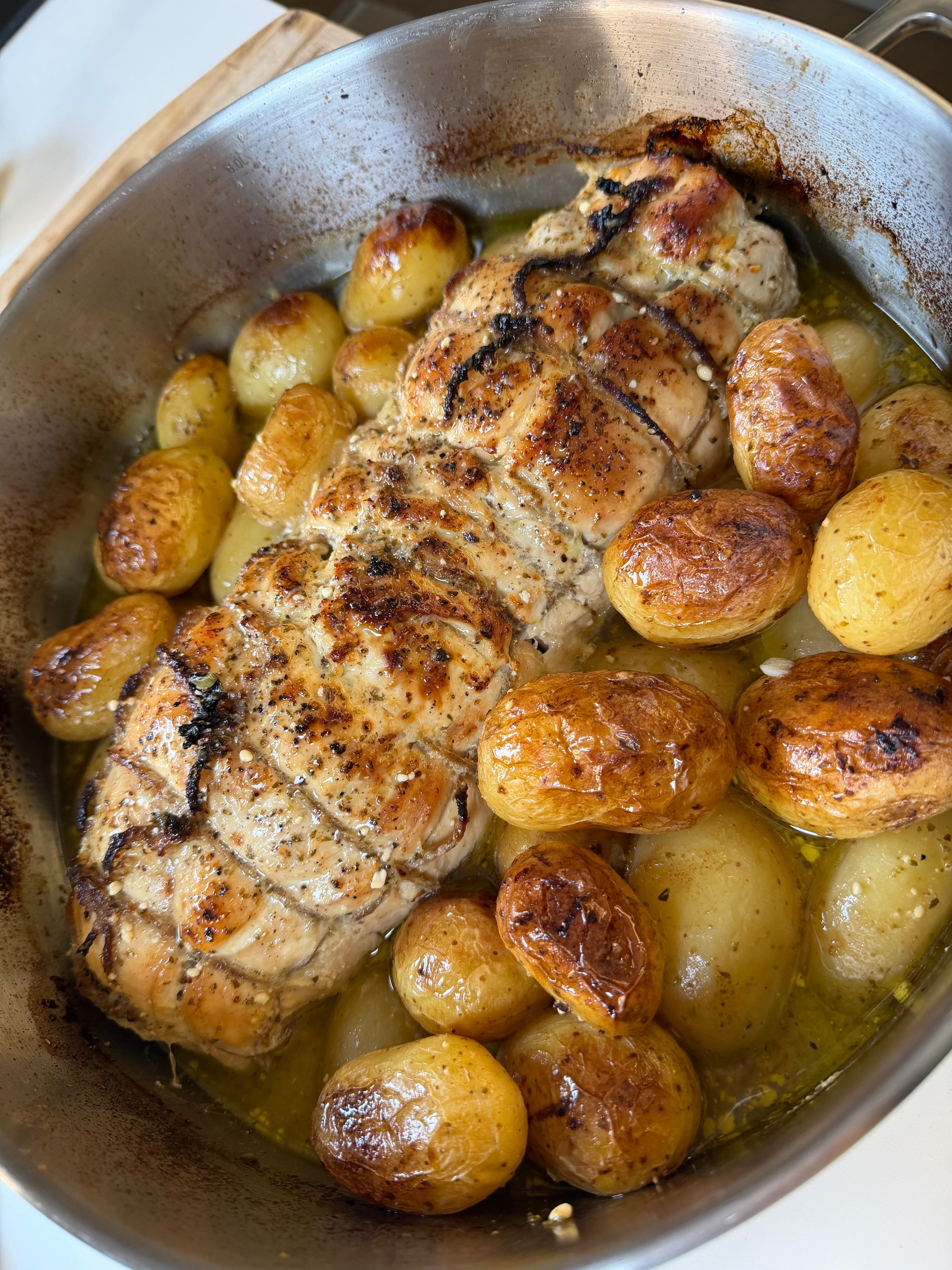 A roasted pork loin tied with string is surrounded by golden-brown roasted potatoes in a metal pan, glistening with oil and herbs.