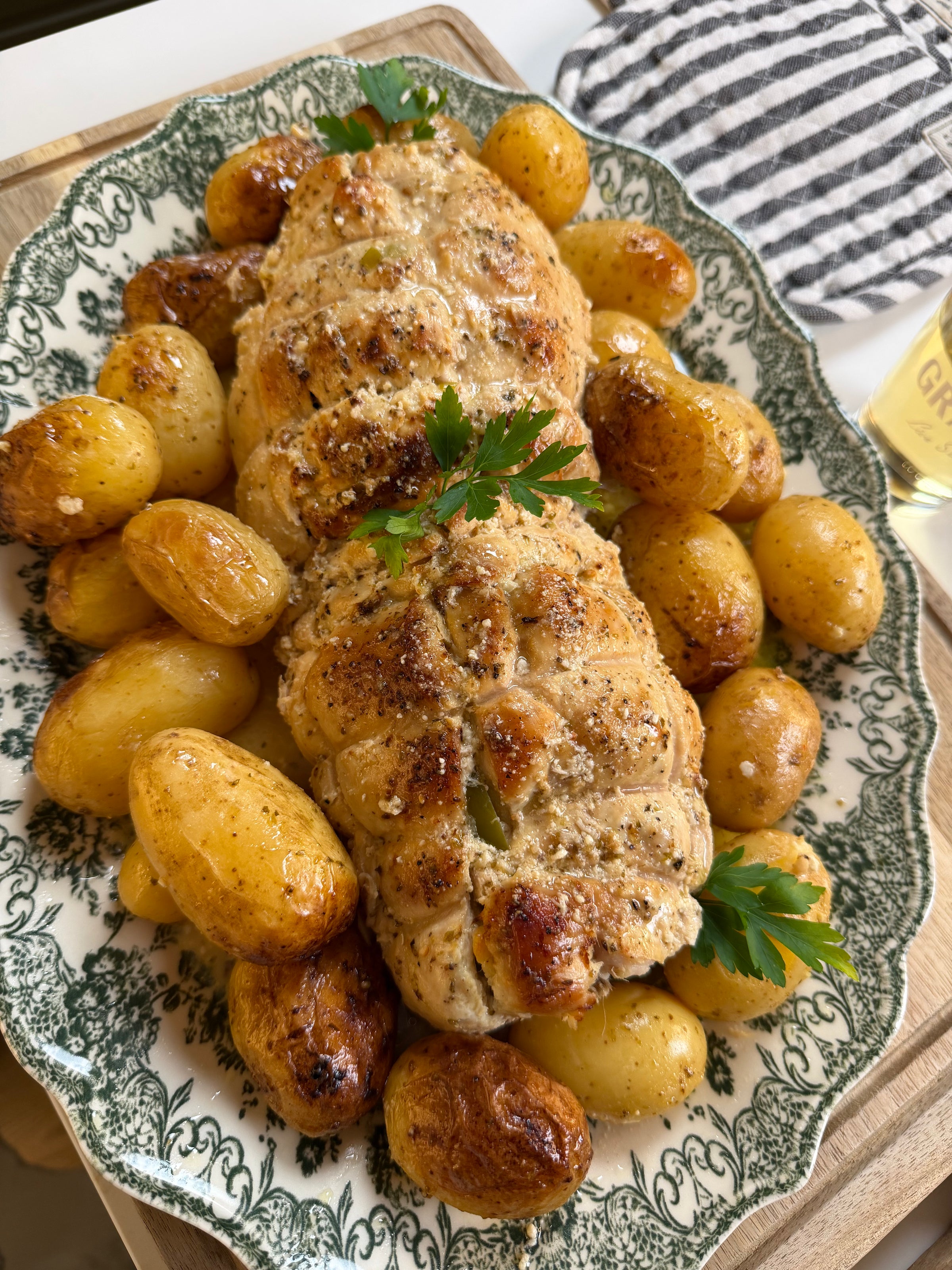 Roasted stuffed chicken breast, tied with twine, is served on a vintage platter with golden roasted baby potatoes and garnished with parsley sprigs. A striped towel and a beverage are visible in the background.