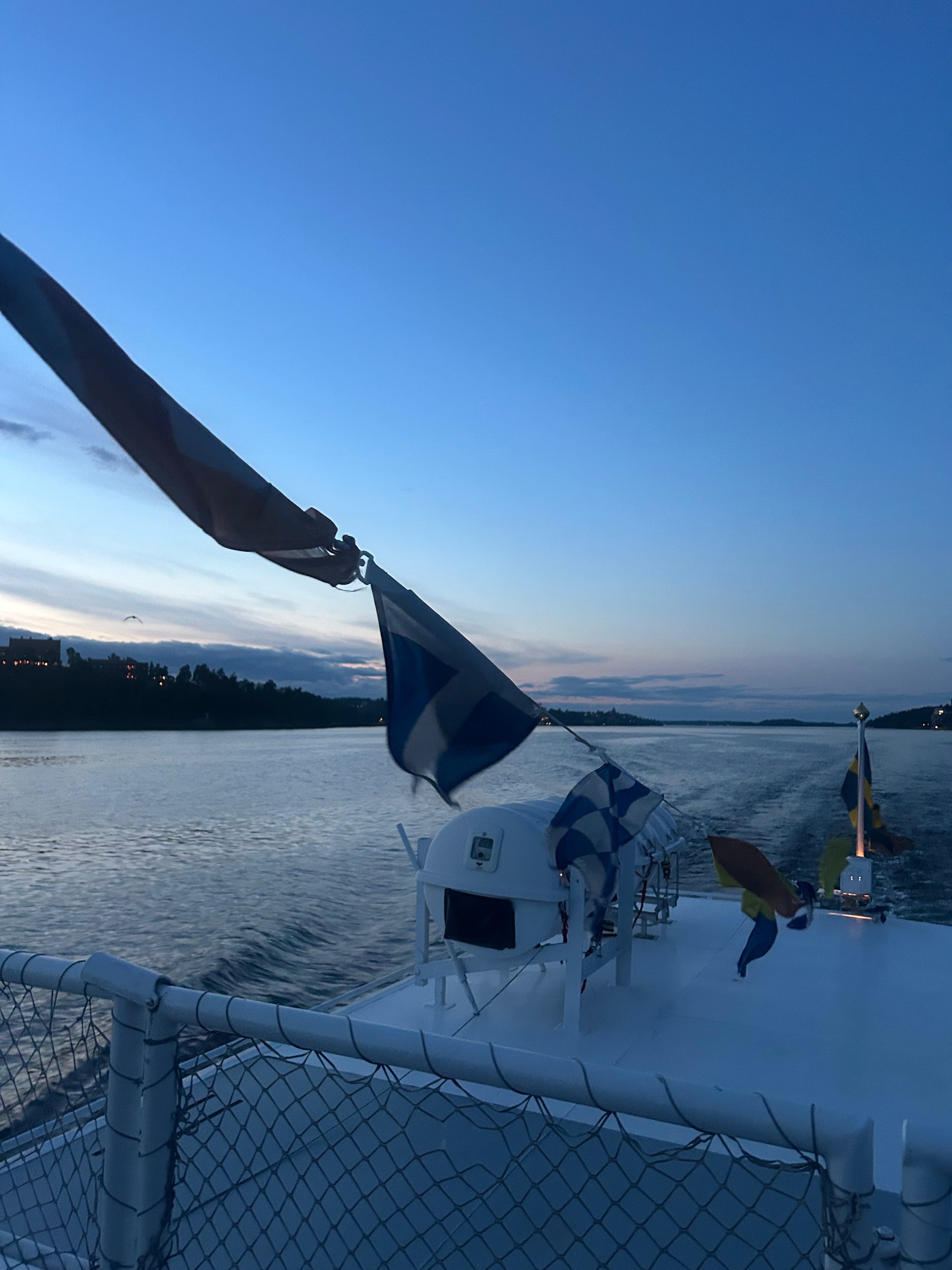 A view from a boat at sunset, with colorful flags strung along the deck. Calm water and a distant shoreline are visible under a blue and orange sky.