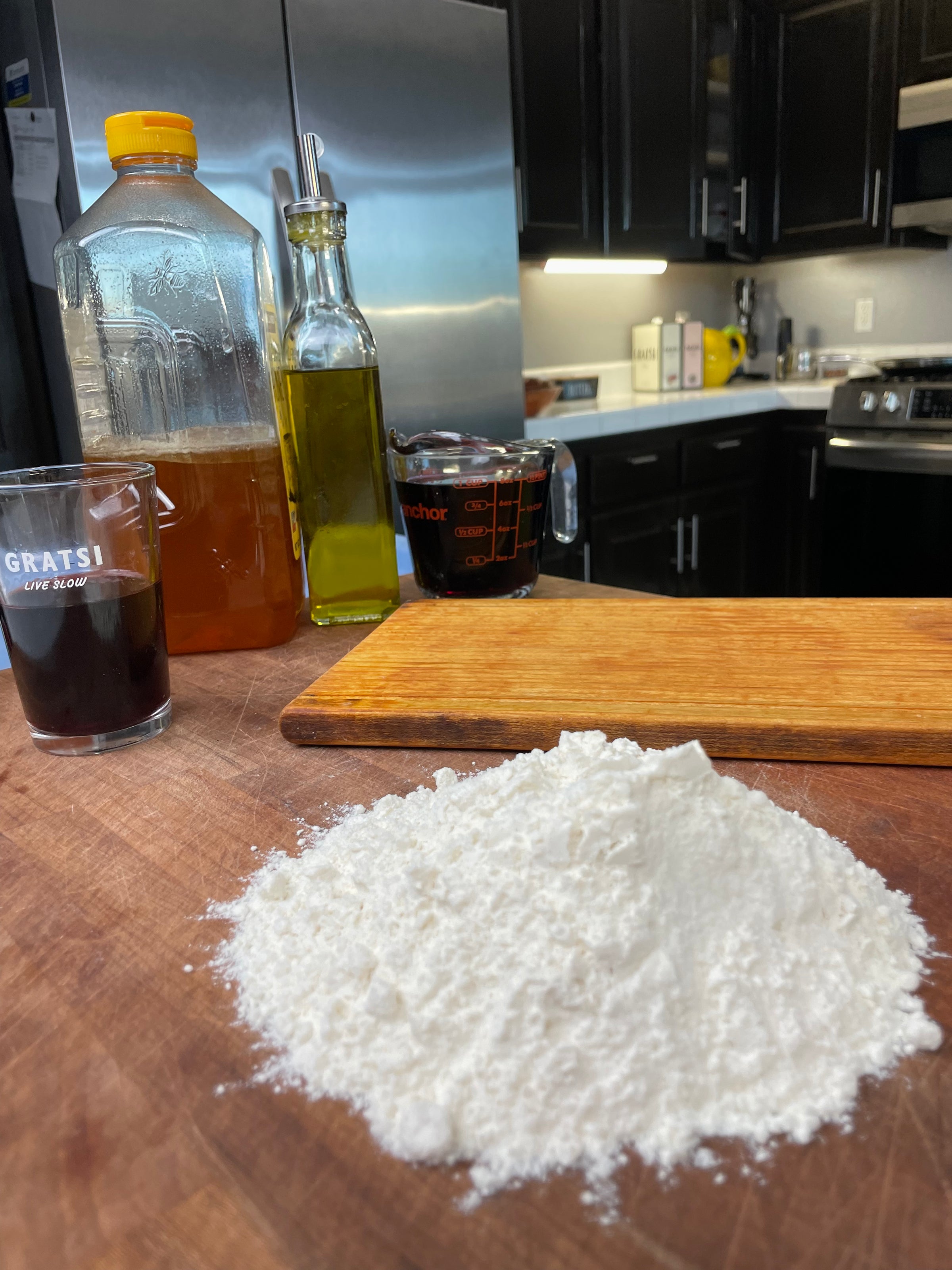 A pile of flour on a wooden cutting board, with olive oil, a glass of dark liquid, a large jug, and a measuring cup in the background in a modern kitchen.