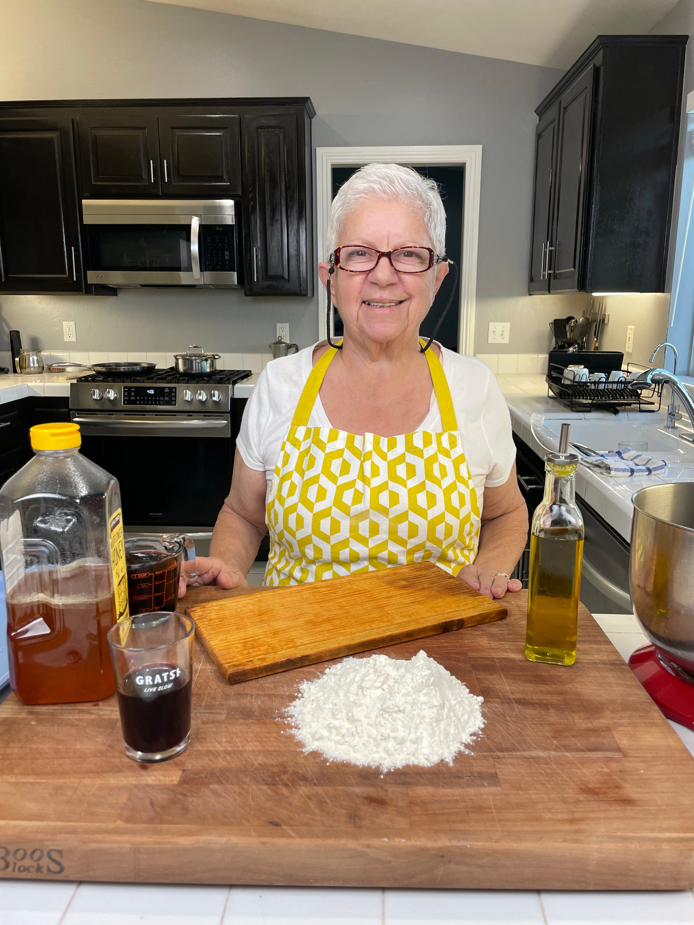 An older woman with short white hair, wearing glasses and a yellow patterned apron, stands in a kitchen behind a wooden counter with flour, olive oil, honey, and a measuring cup on it. She is smiling at the camera.