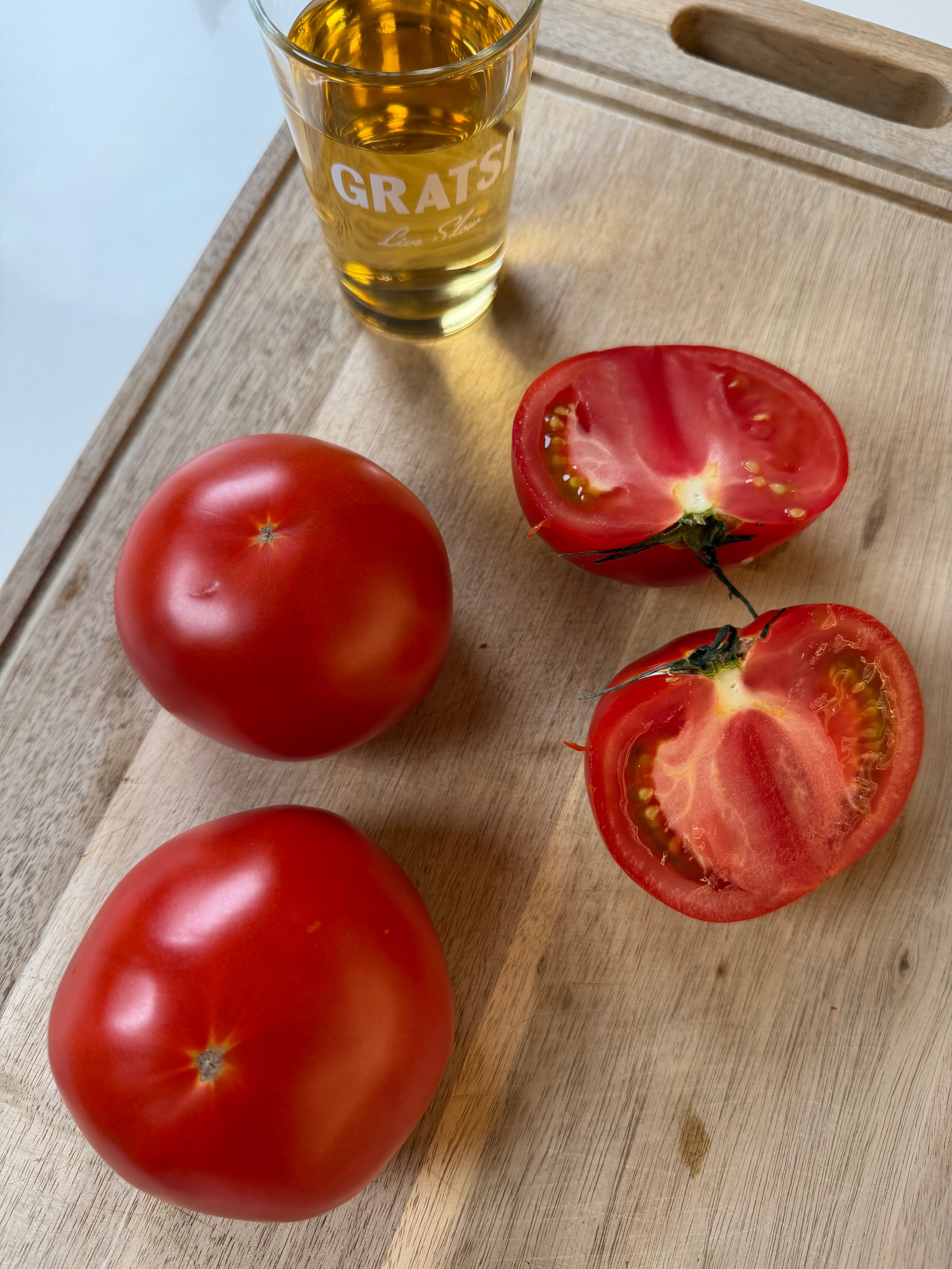 Two whole tomatoes and one halved tomato on a wooden cutting board, next to a small glass of yellow liquid on a white surface.