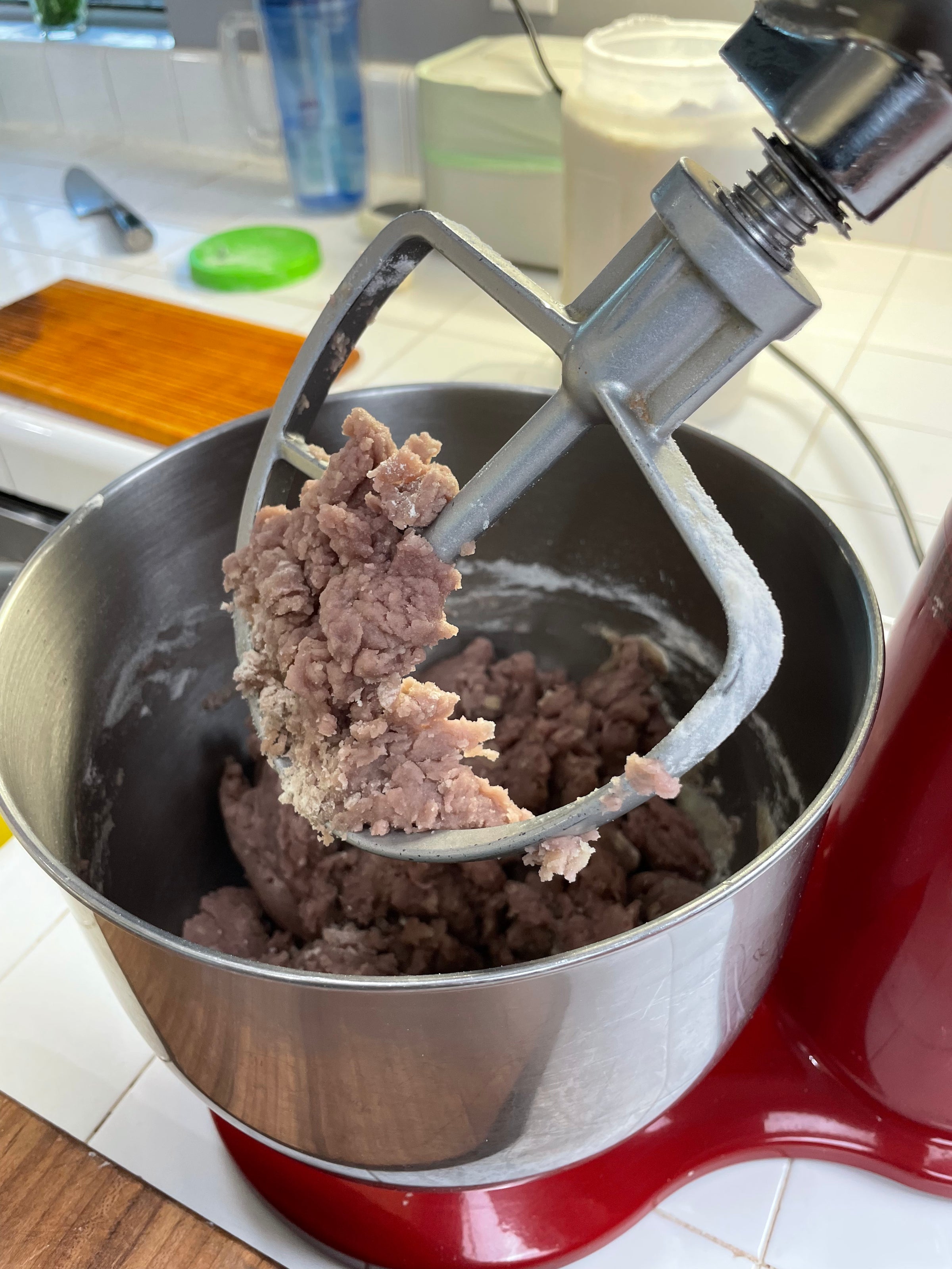 A close-up of a stand mixer with a paddle attachment mixing a thick, light purple dough in a metal bowl on a kitchen counter. Various kitchen items are visible in the background.