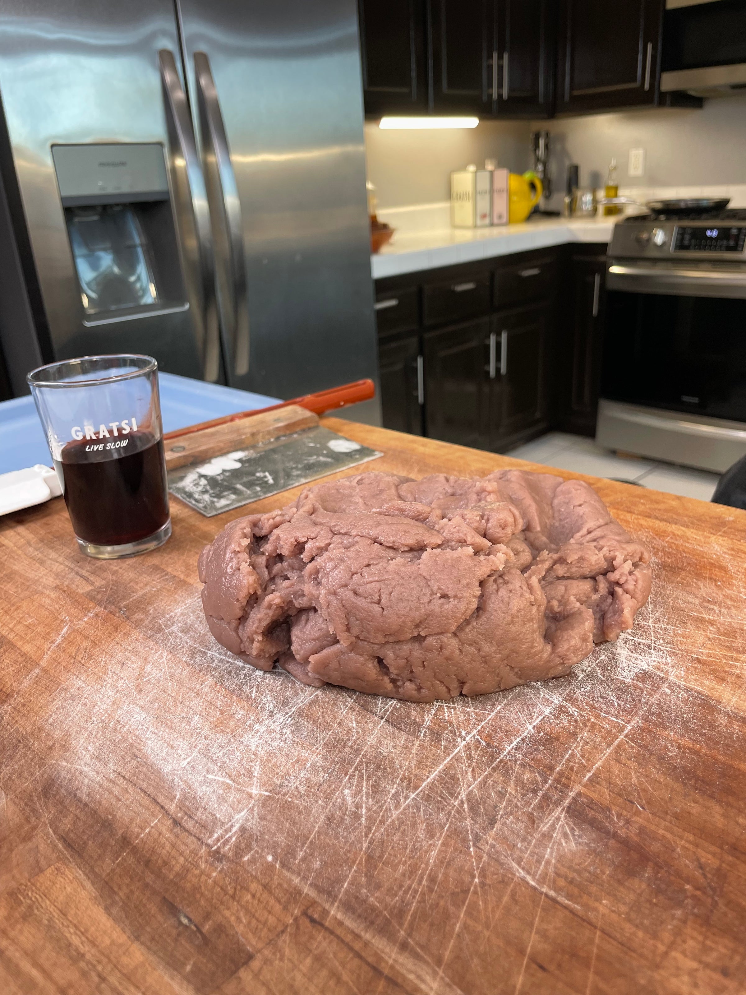 A ball of purple dough rests on a floured wooden countertop in a modern kitchen, with a glass of dark liquid, bench scraper, and kitchen appliances visible in the background.