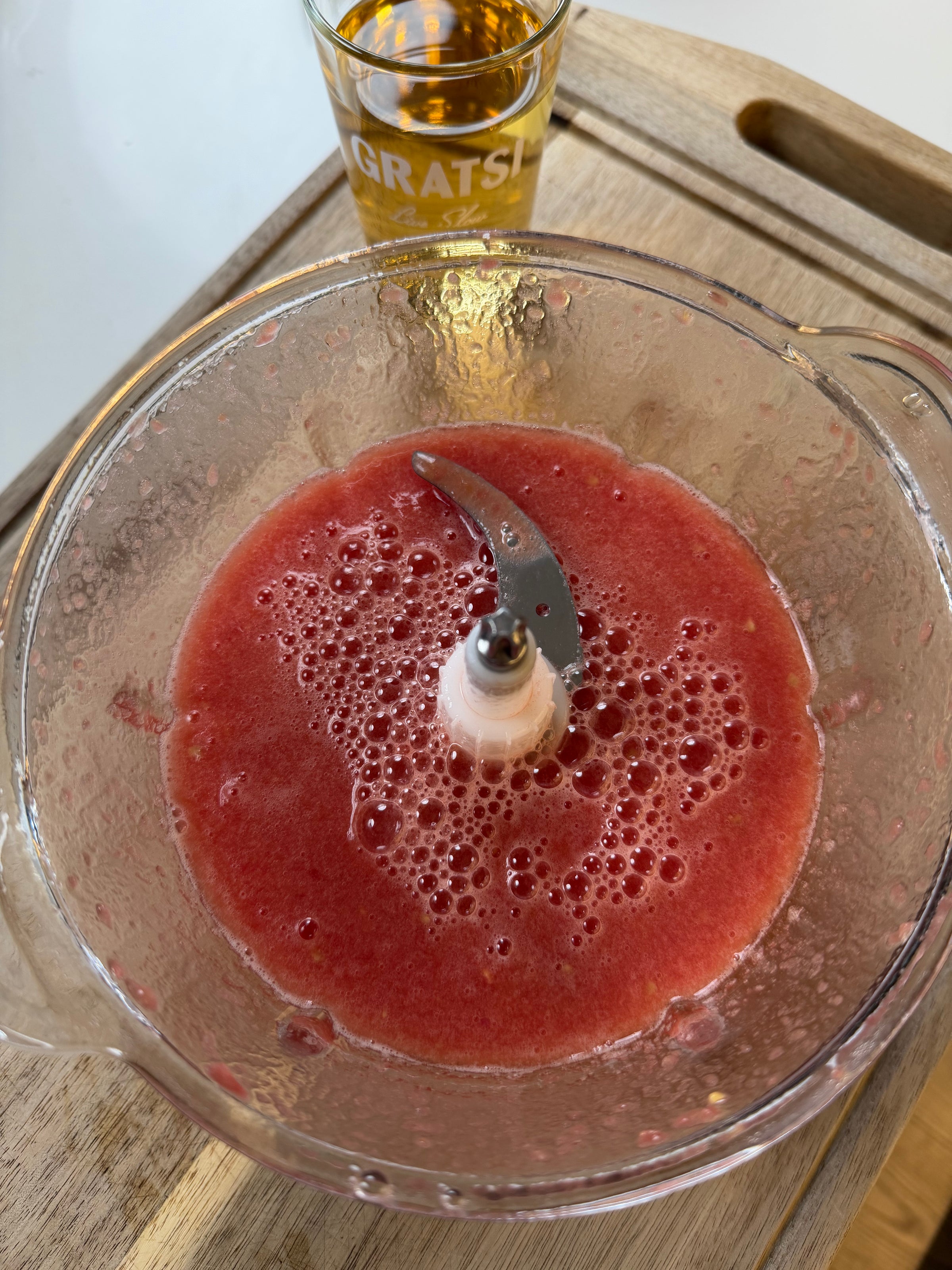 A food processor bowl filled with blended pink watermelon sits on a wooden surface. Behind it, a glass with yellow liquid and the word GRATSI is partially visible.
