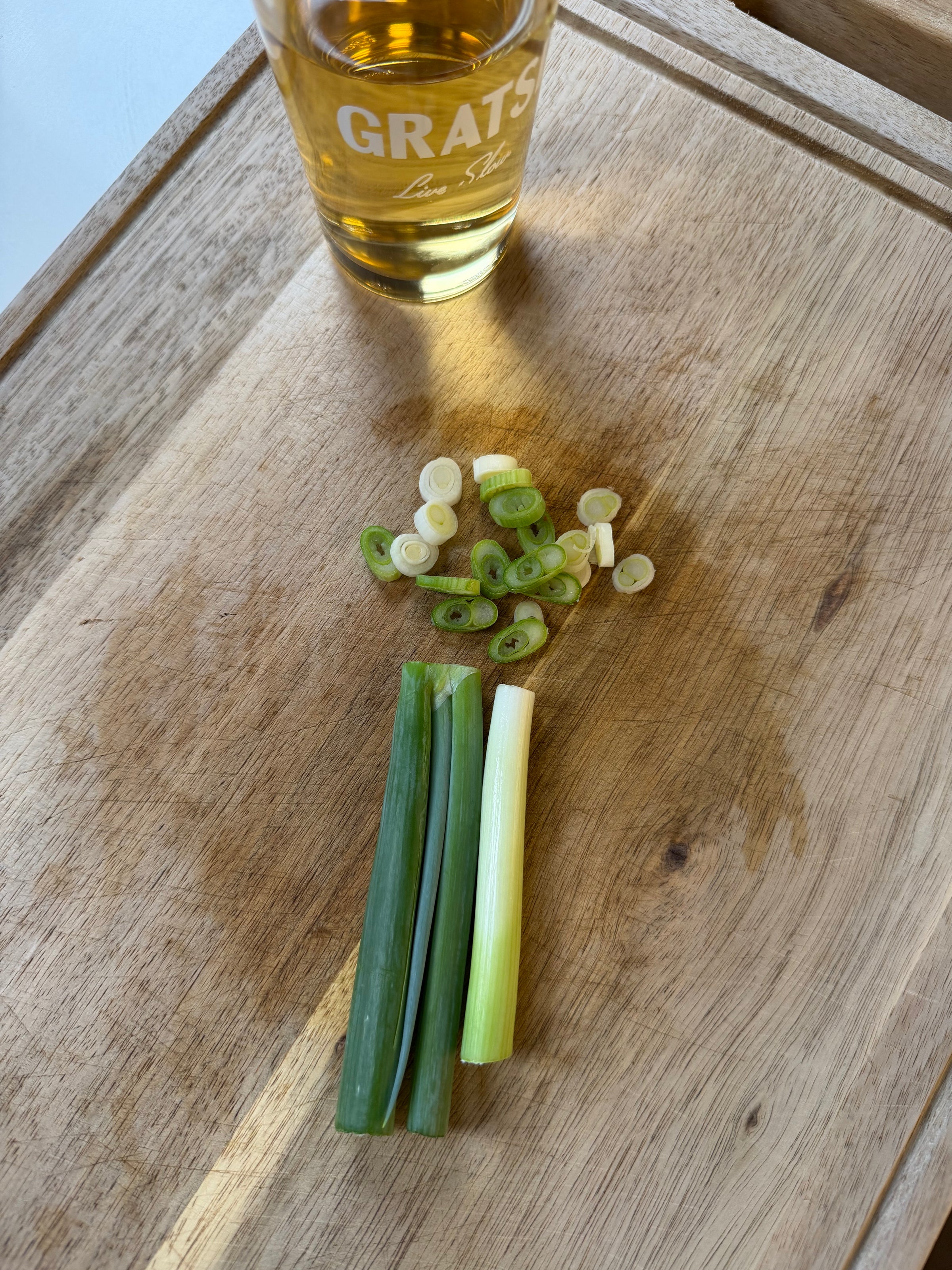 A glass of amber liquid sits on a wooden surface next to chopped and whole green onions, neatly arranged on a cutting board.