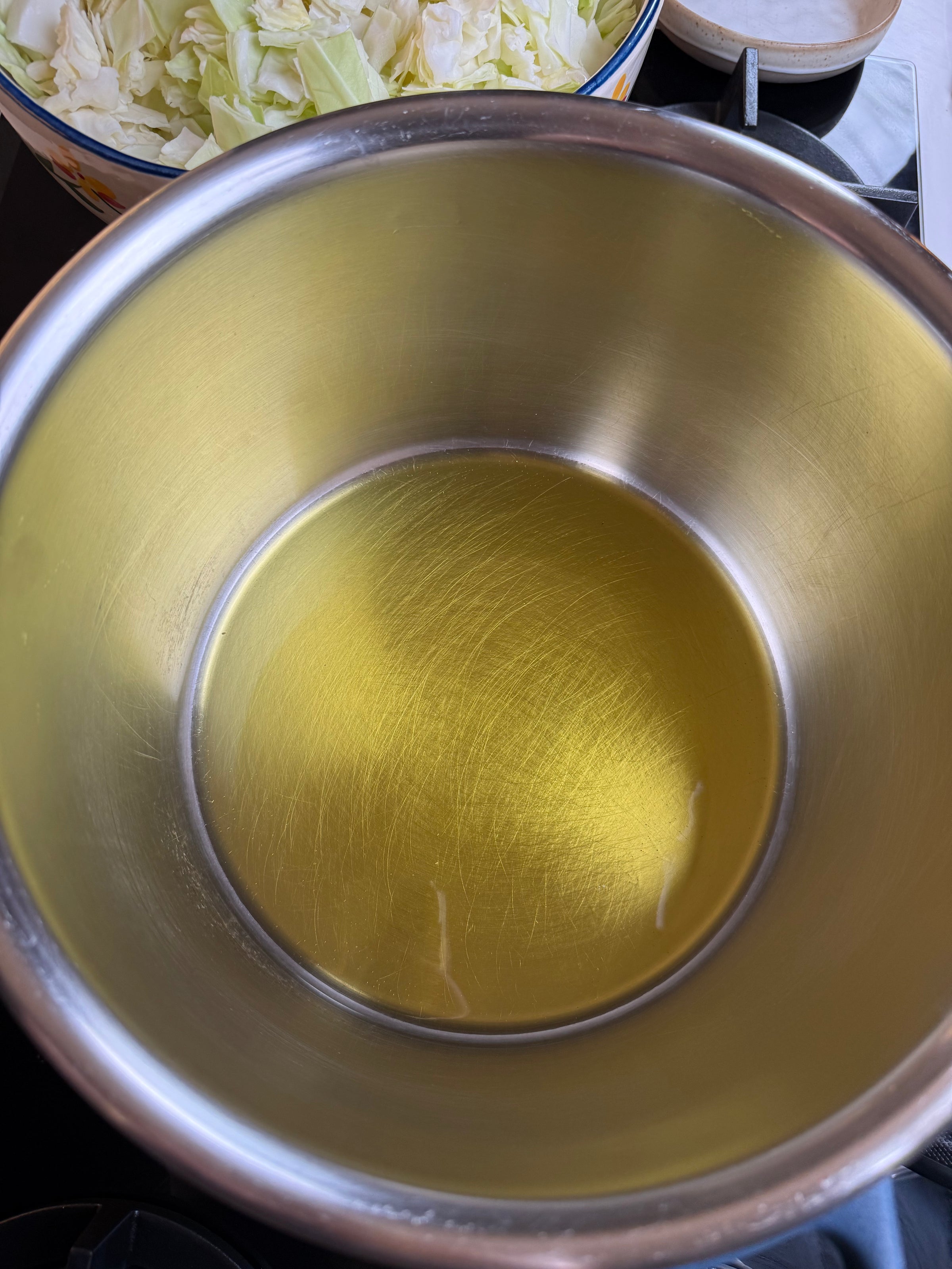A stainless steel pot with a thin layer of oil at the bottom sits on a stove. In the background, there is a bowl filled with chopped cabbage.