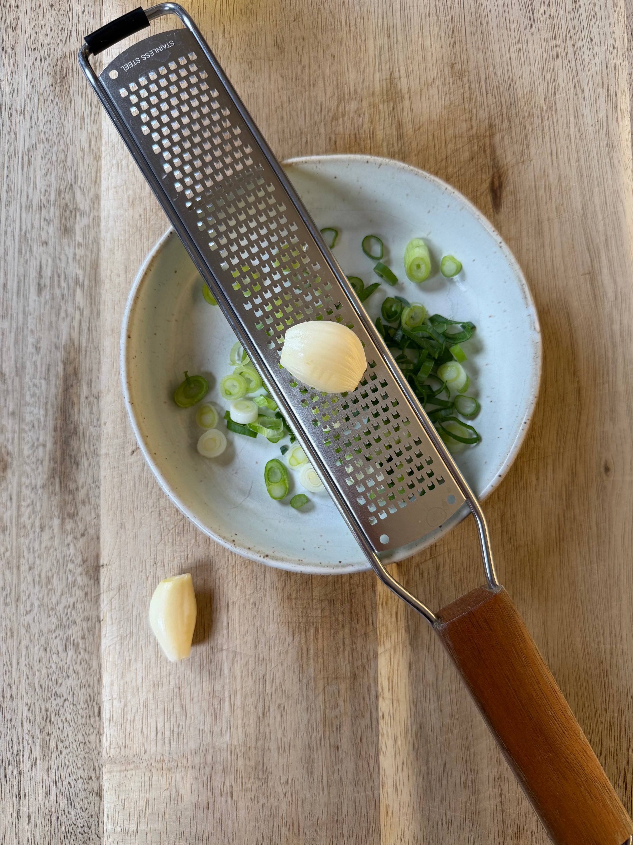 A garlic clove and a grater rest on a bowl of sliced green onions, all on a wooden surface. Another garlic clove sits nearby on the cutting board.