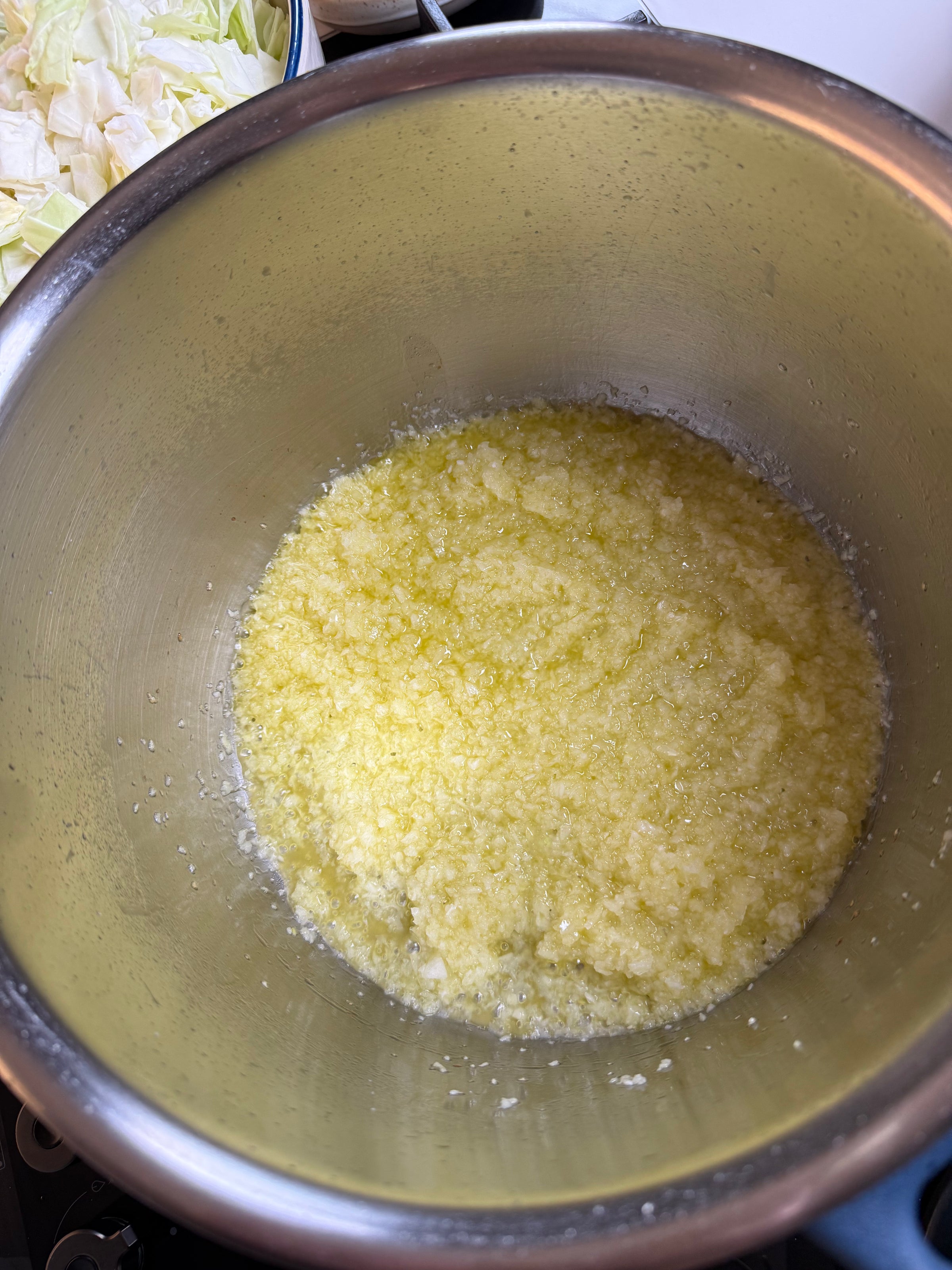 A large metal mixing bowl filled with finely grated or minced garlic mixed with oil, creating a yellowish, textured mixture. Some chopped cabbage is visible in the corner of the image.