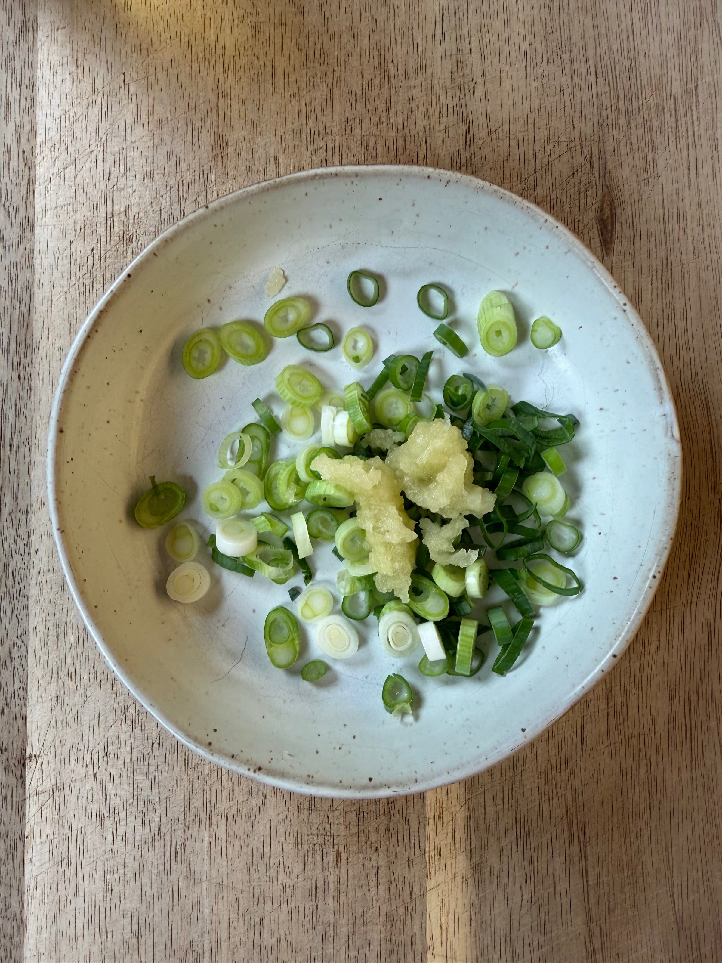 A white plate on a wooden surface holds chopped green onions and a small pile of minced garlic.