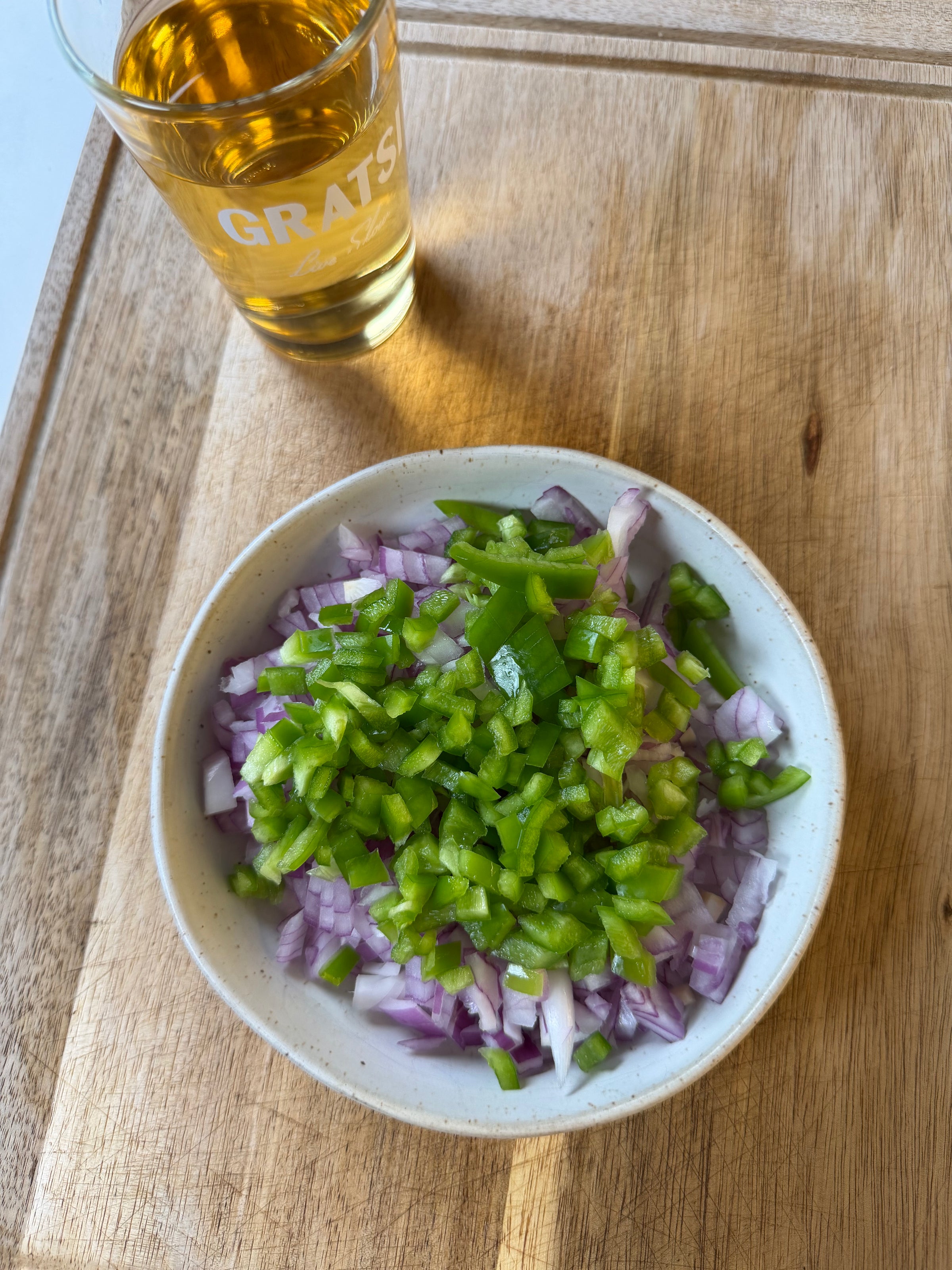 A bowl filled with chopped red onions and green bell peppers sits on a wooden surface next to a glass of amber-colored liquid.