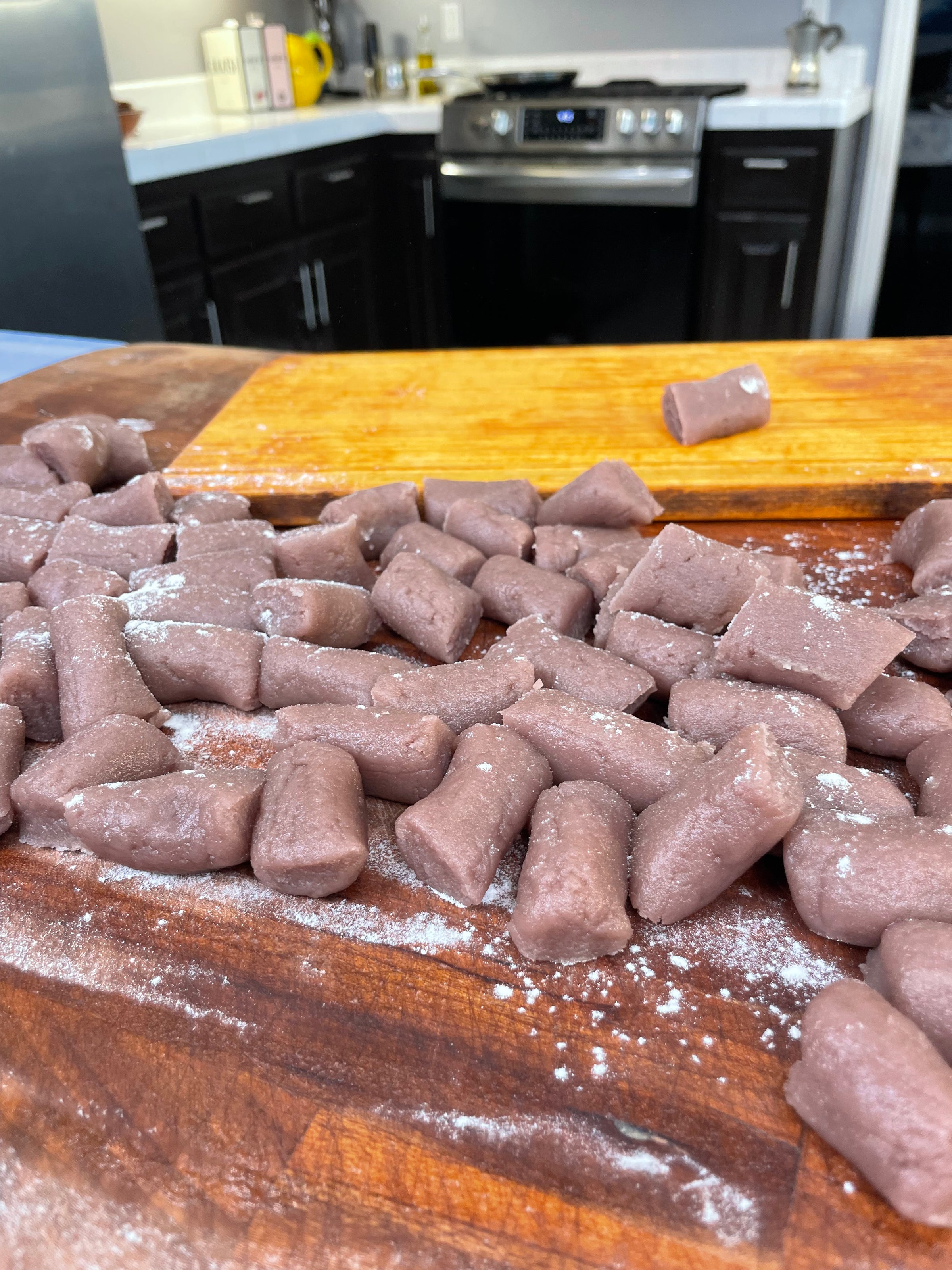 A pile of uncooked purple gnocchi pieces dusted with flour sits on a wooden surface in a kitchen, with a stove, cabinets, and countertop visible in the background.