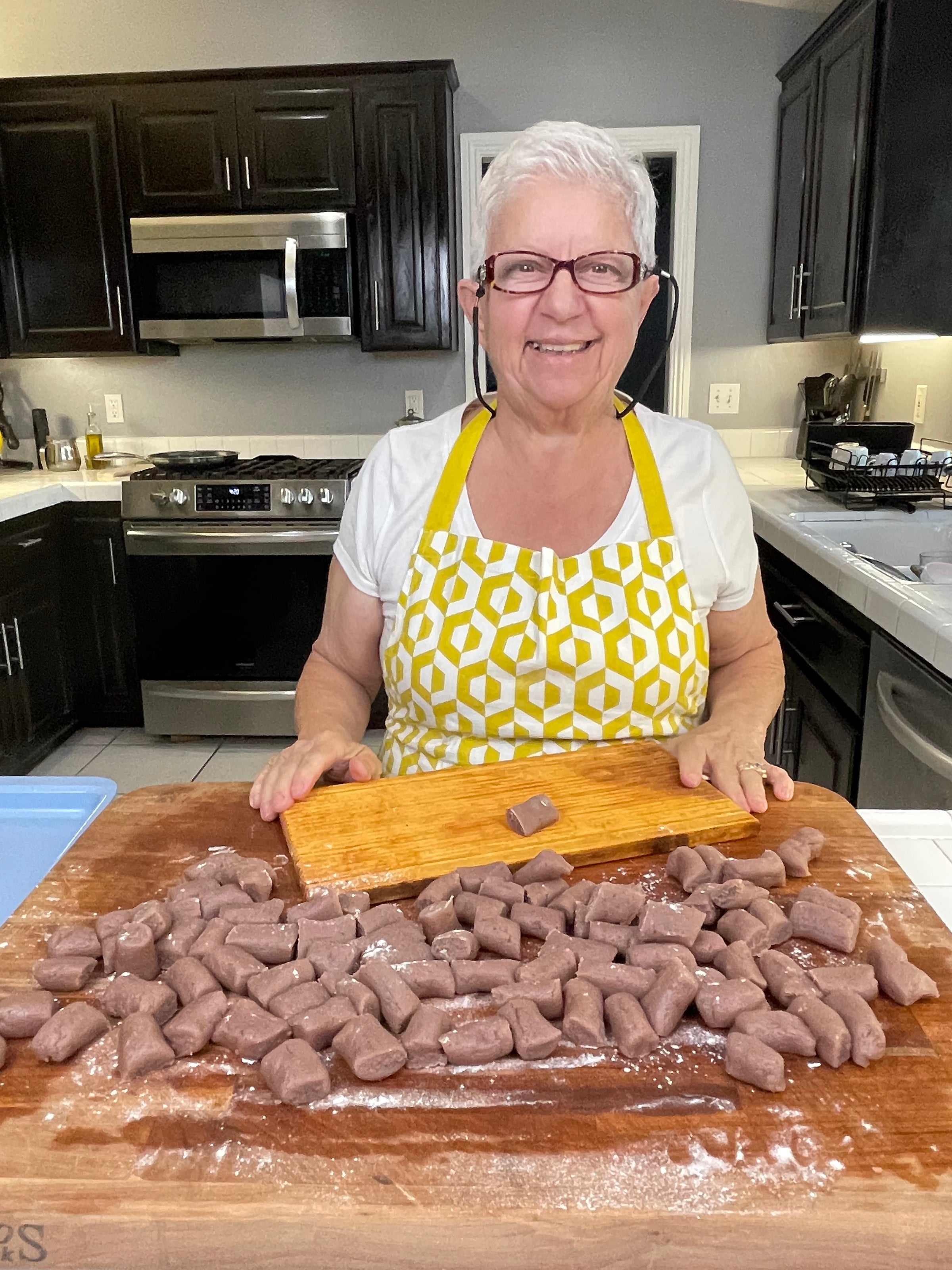 Smiling elderly woman with short white hair and glasses stands in a modern kitchen, wearing a yellow patterned apron, holding a cutting board behind a counter covered with pieces of uncooked dough.