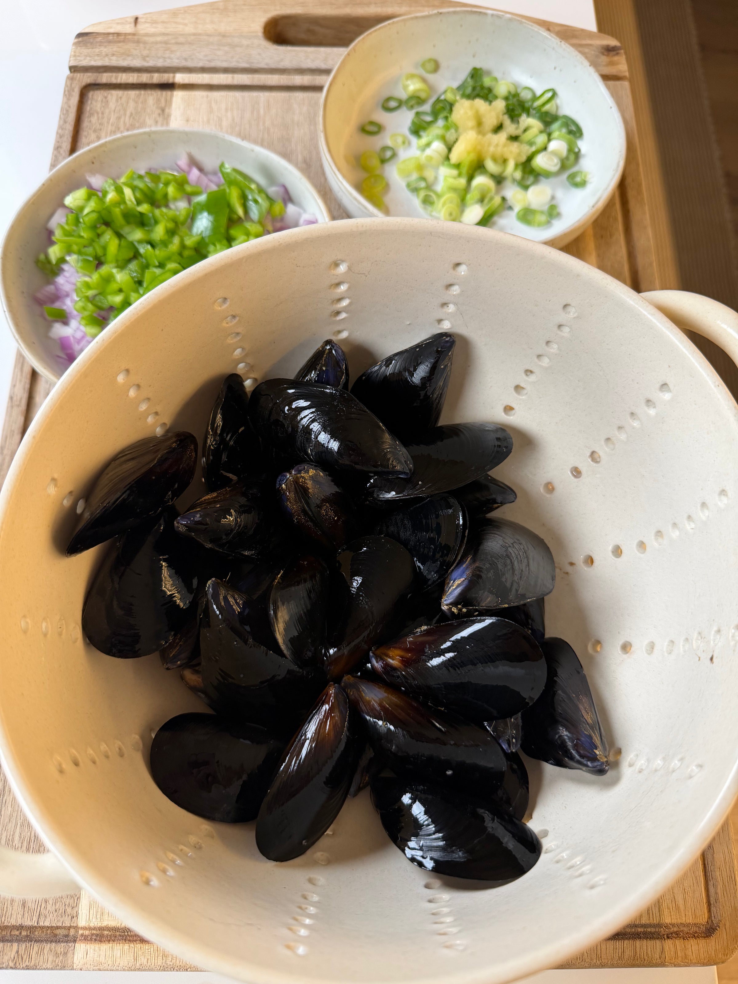 A white colander with black mussels sits on a wooden board, next to bowls of chopped green onions and green peppers in a light sauce, ready for cooking.