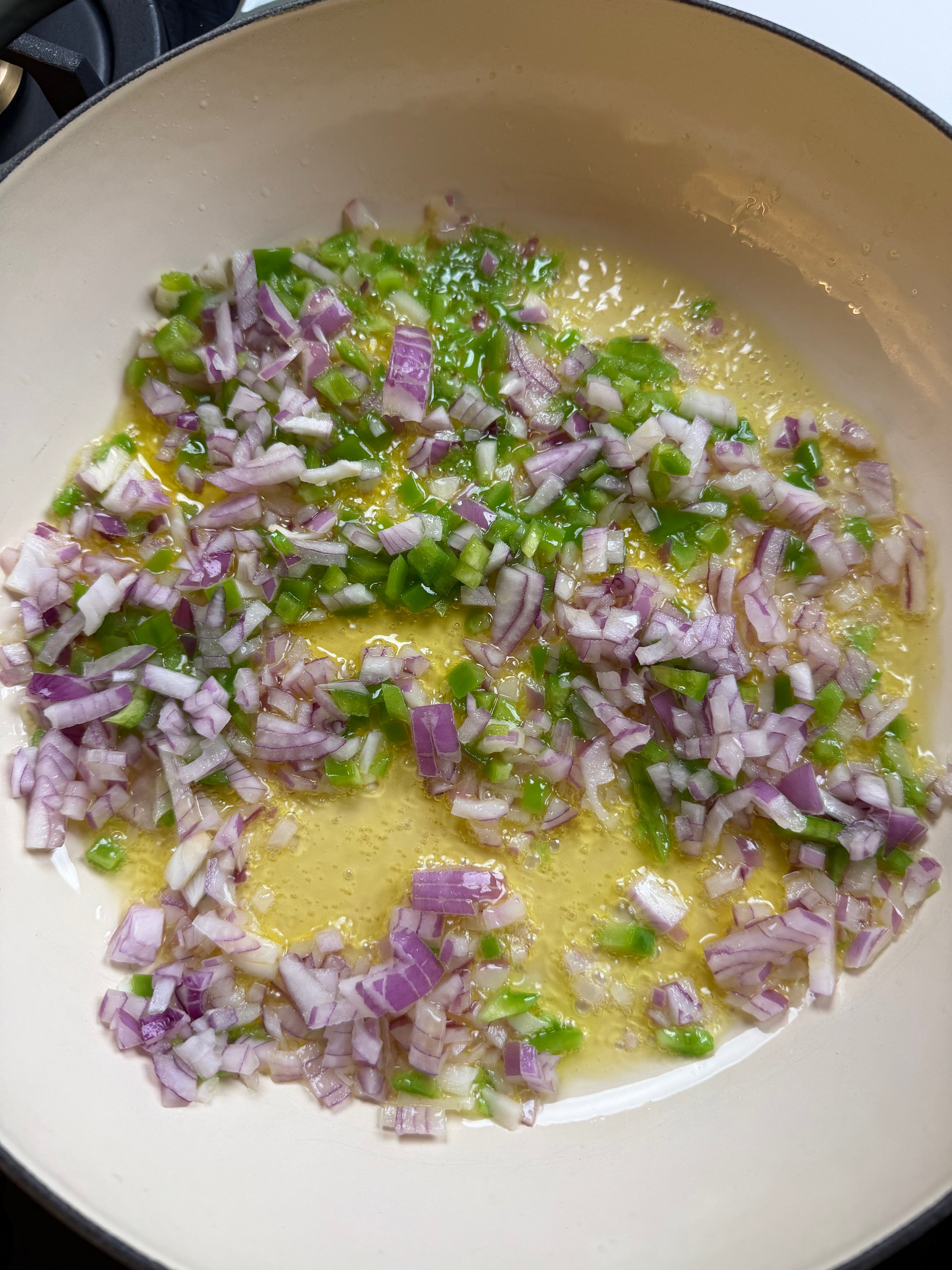 Chopped red onions and green bell peppers are being sautéed in oil in a white pan on a stovetop.