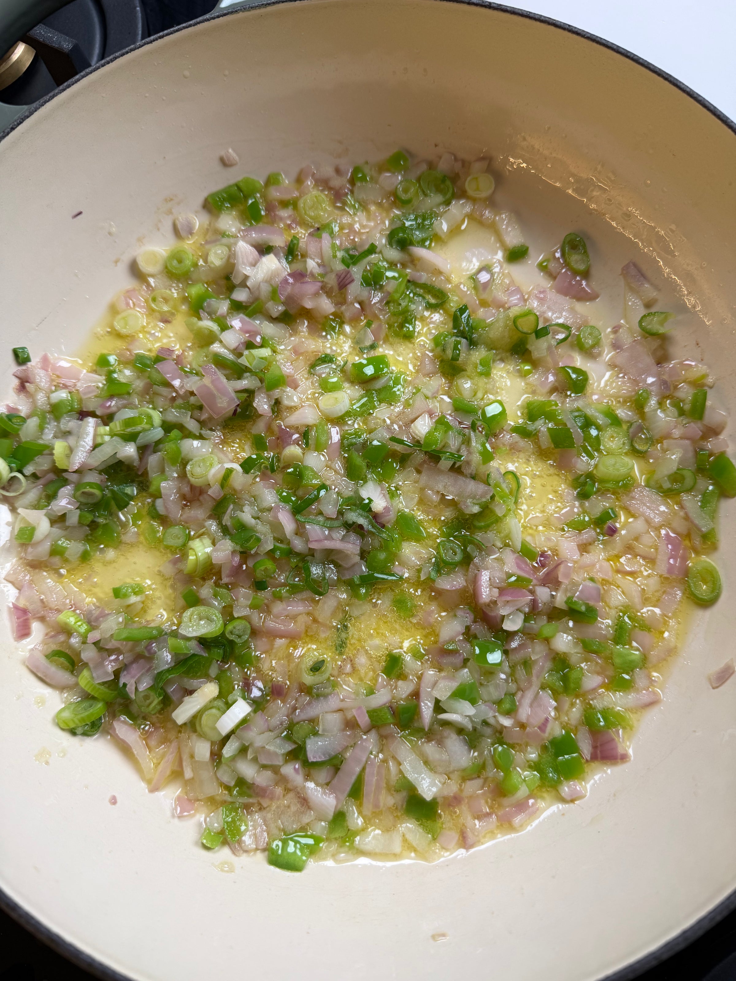 Chopped green peppers, onions, and shallots are being sautéed in oil in a light-colored pan.