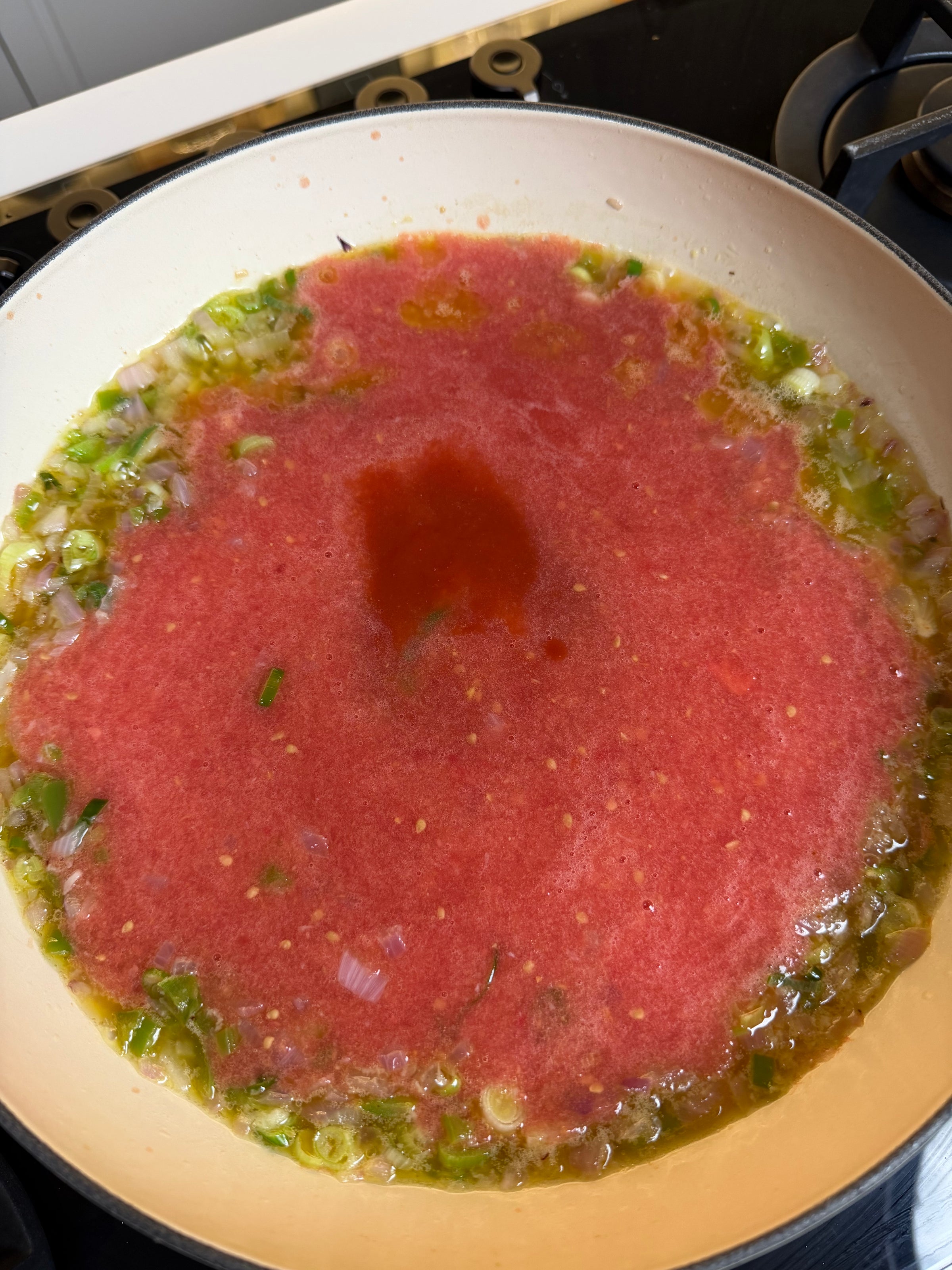 A pan on a stove contains chopped green onions and onions sautéing, with a large amount of blended tomato sauce added in the center, starting to simmer.