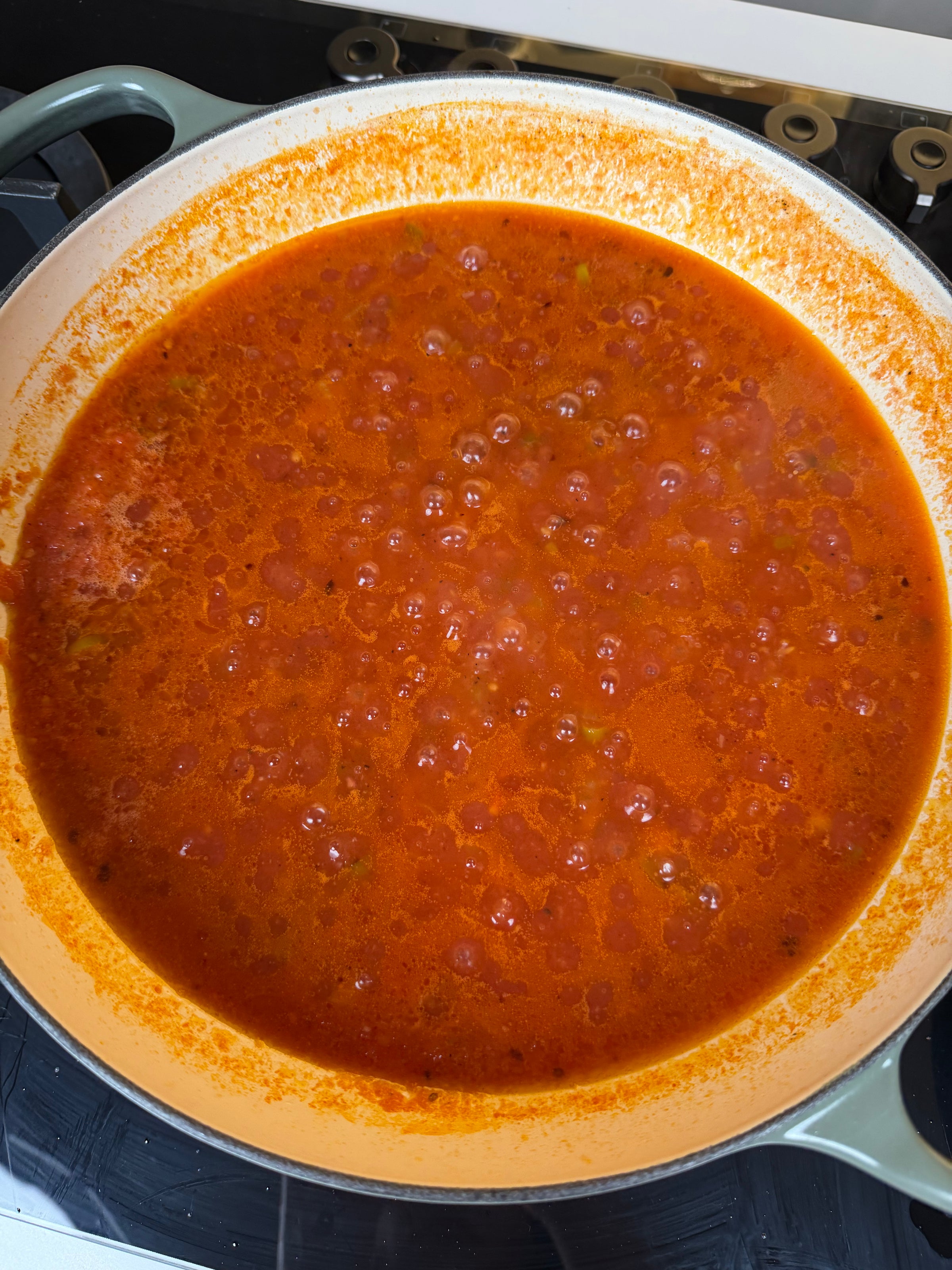 A large pot filled with bubbling red tomato sauce simmers on a stovetop, with orange oil pooling around the edges and visible bubbles breaking the surface.