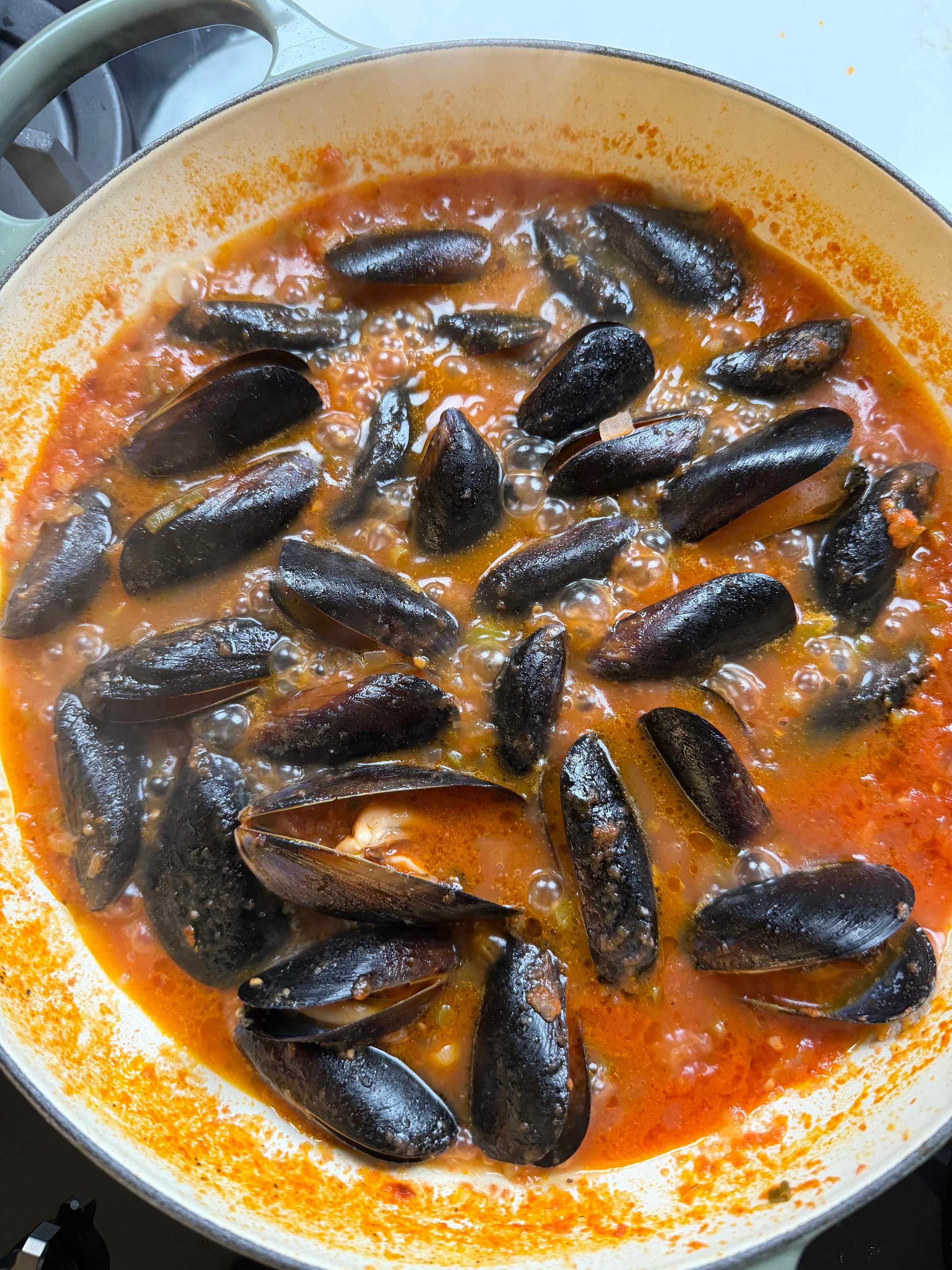 A pot filled with black mussels simmering in a red tomato-based sauce, with bubbles rising as the dish cooks on the stove.