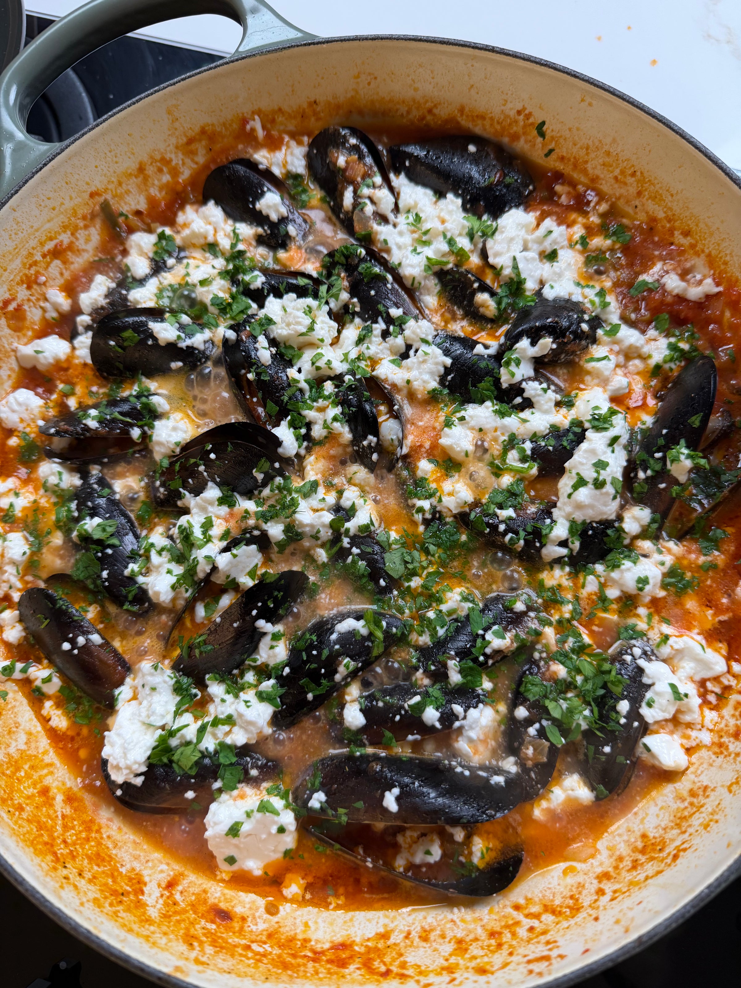 A pot of mussels cooked in tomato sauce, topped with crumbled feta cheese and chopped fresh parsley, sits on a stovetop.