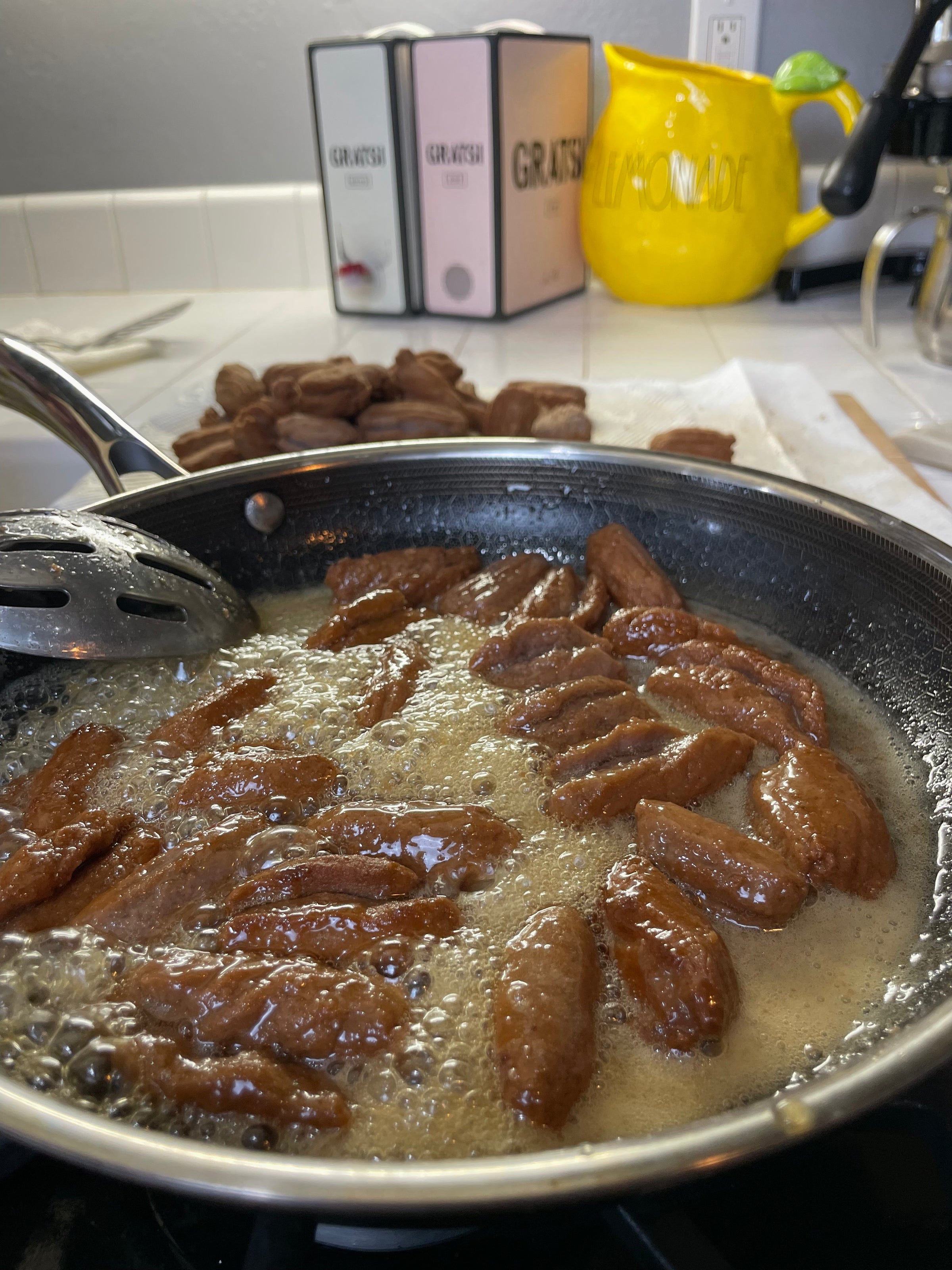A frying pan filled with sizzling, brown dough pieces in bubbling oil on a stove. In the background, a stack of finished pastries, three labeled canisters, and a yellow lemon-shaped container are visible.