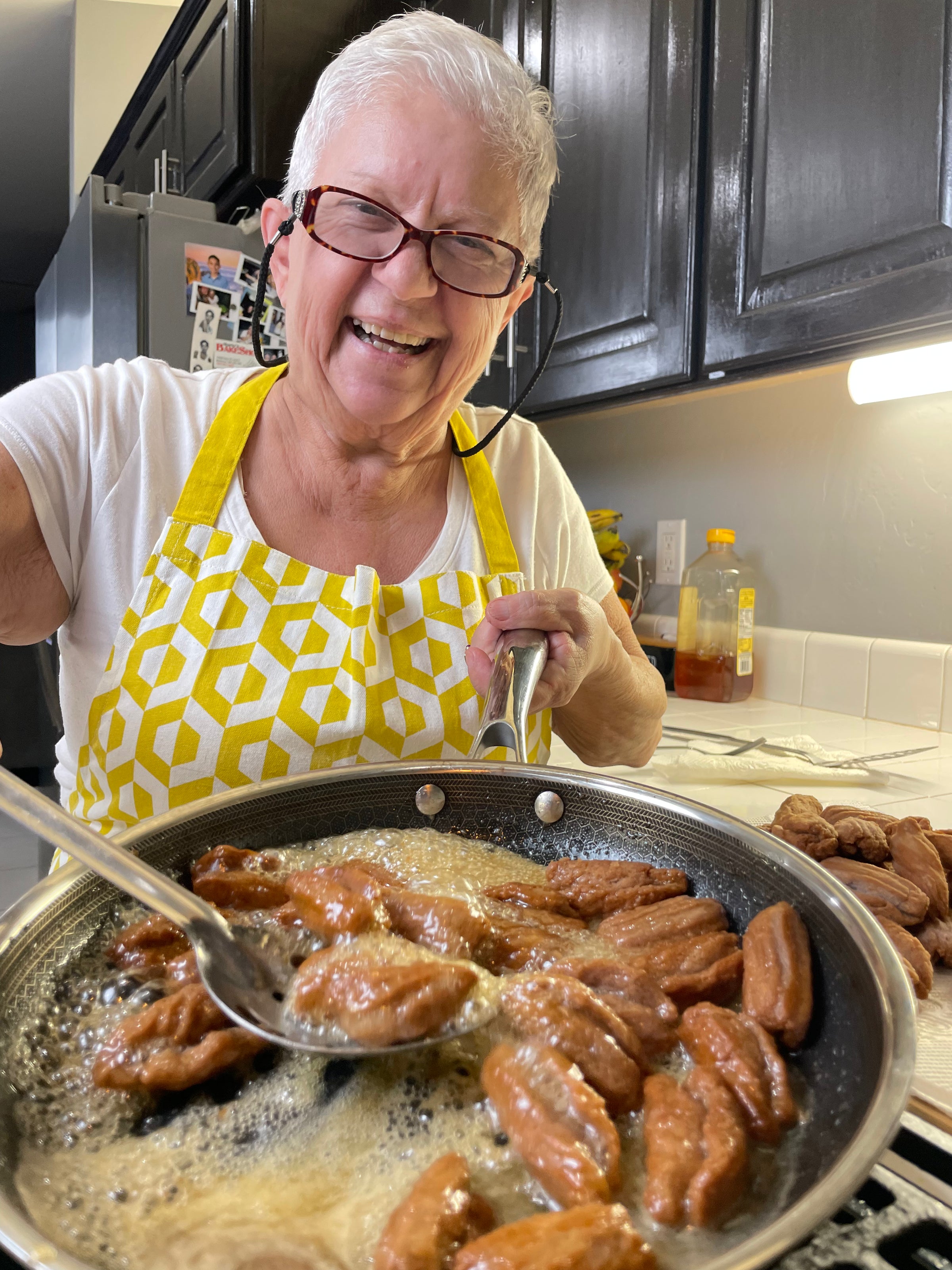 An older woman with short white hair and glasses, wearing a yellow patterned apron, smiles while frying pastries in a pan on a stove in a modern kitchen.