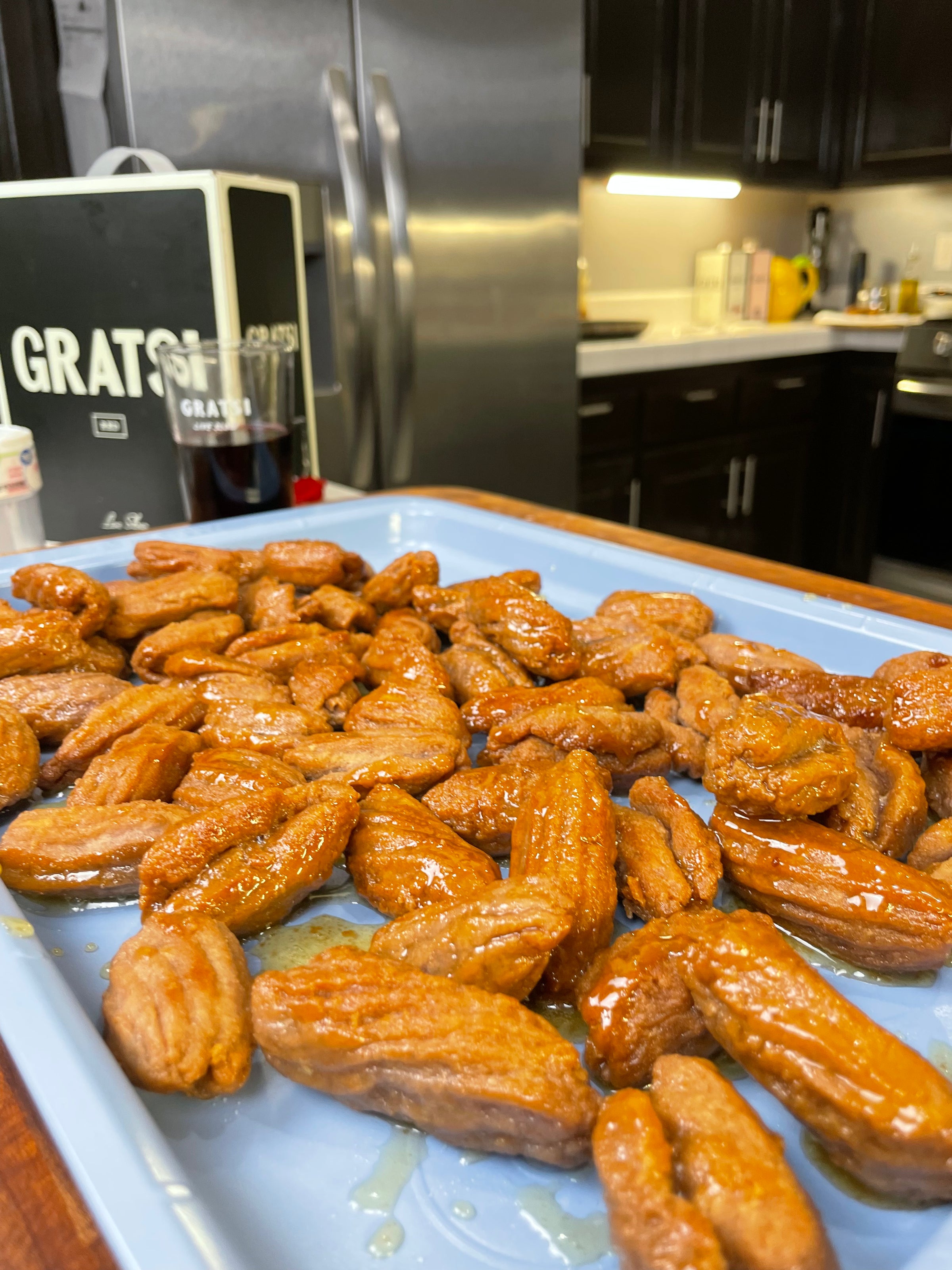 A tray of golden, honey-coated Italian pastries sits on a blue tray in a modern kitchen with dark cabinets and stainless steel appliances in the background.
