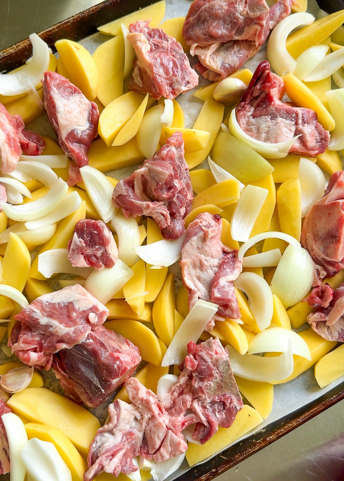Raw pieces of meat, sliced yellow potatoes, and chopped white onions arranged on a baking sheet, ready to be cooked.
