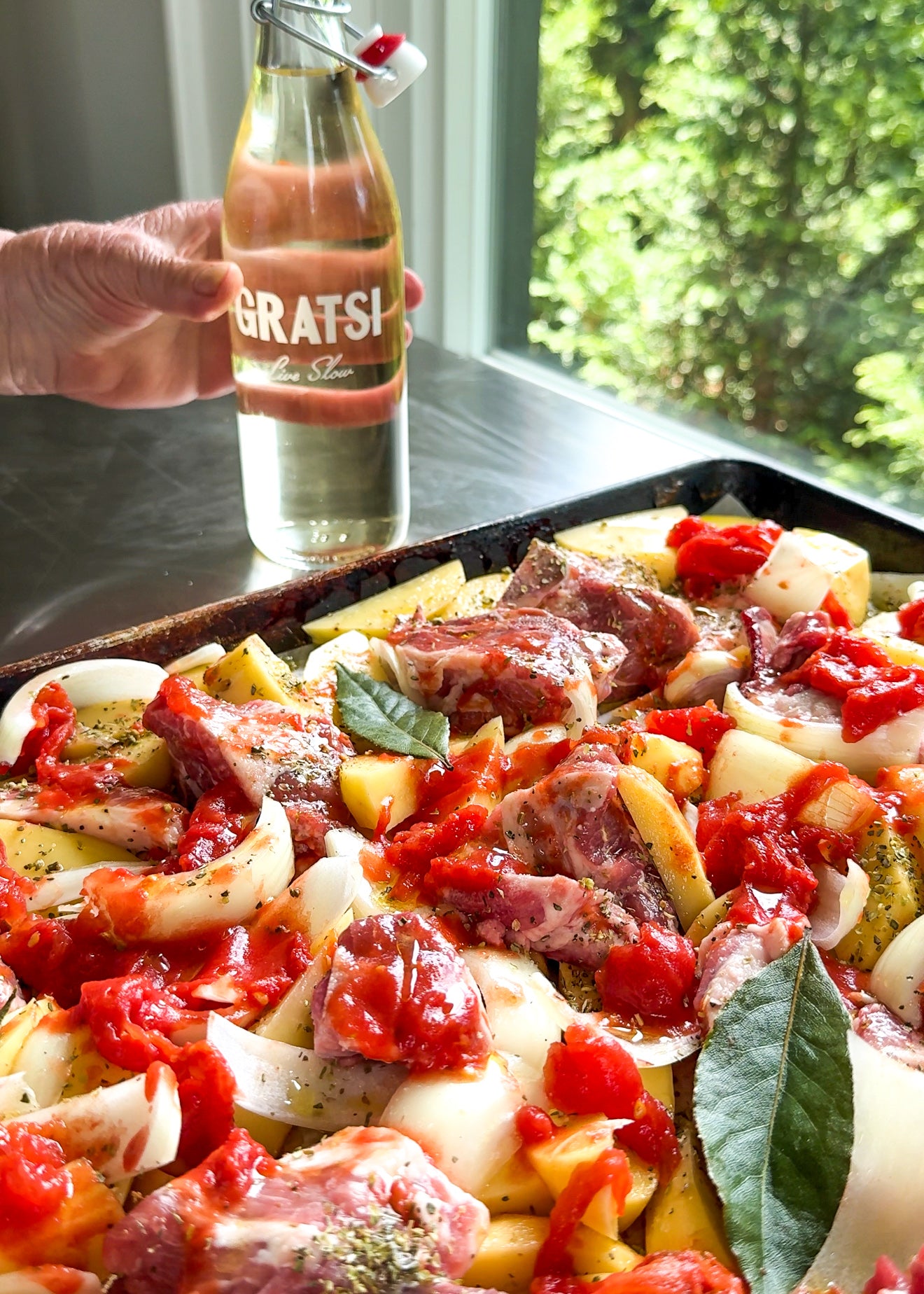 A hand holds a bottle labeled Gratsi near a tray filled with raw meat, onions, potatoes, tomatoes, and bay leaves, set by a window with green foliage outside.