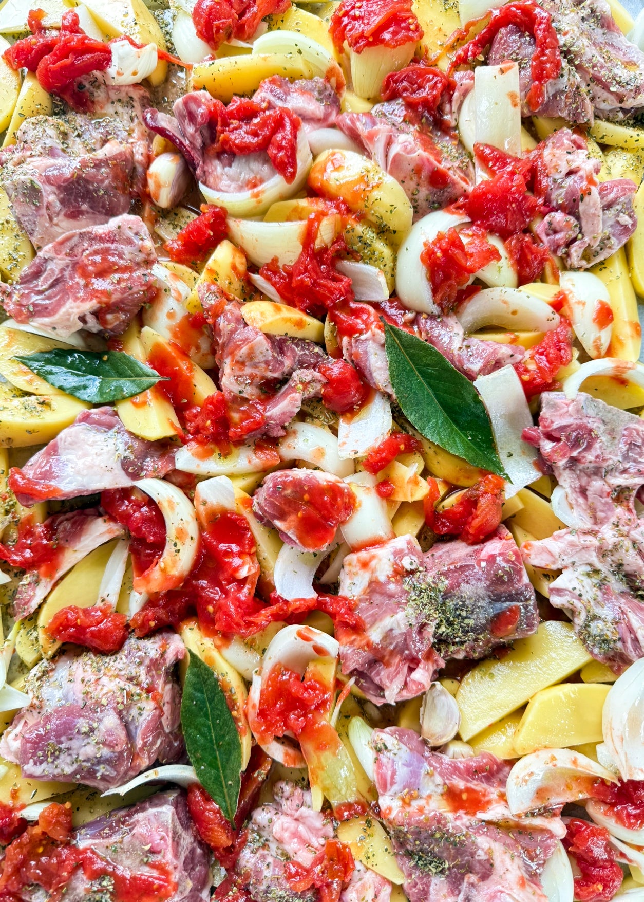 Raw ingredients for a stew, including chunks of meat, sliced onions, potato wedges, bay leaves, diced tomatoes, and seasonings, arranged together before cooking.