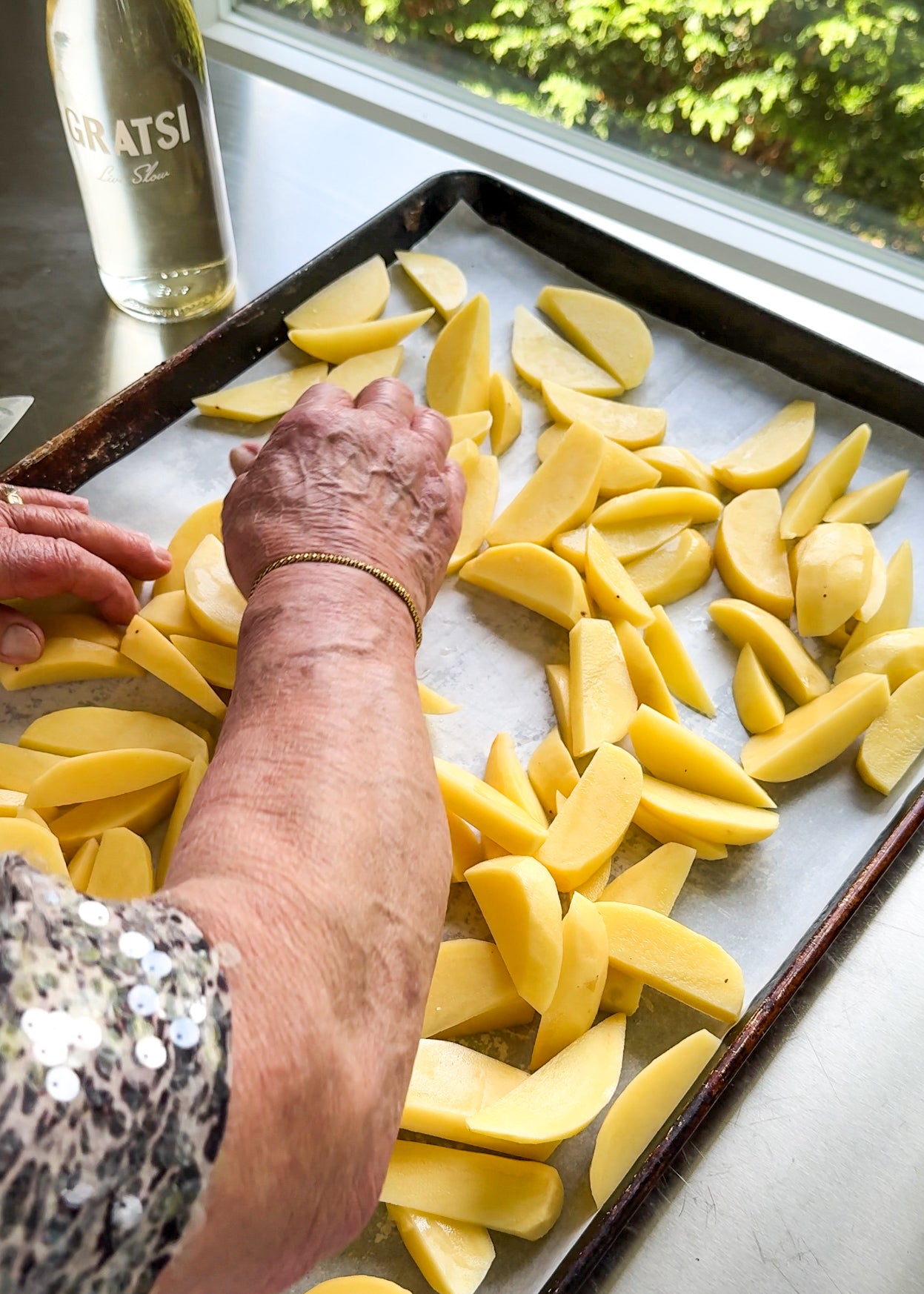 An elderly person arranges raw potato wedges on a baking tray lined with parchment paper, near a window with greenery outside. A bottle labeled GRATSI sits on the counter nearby.