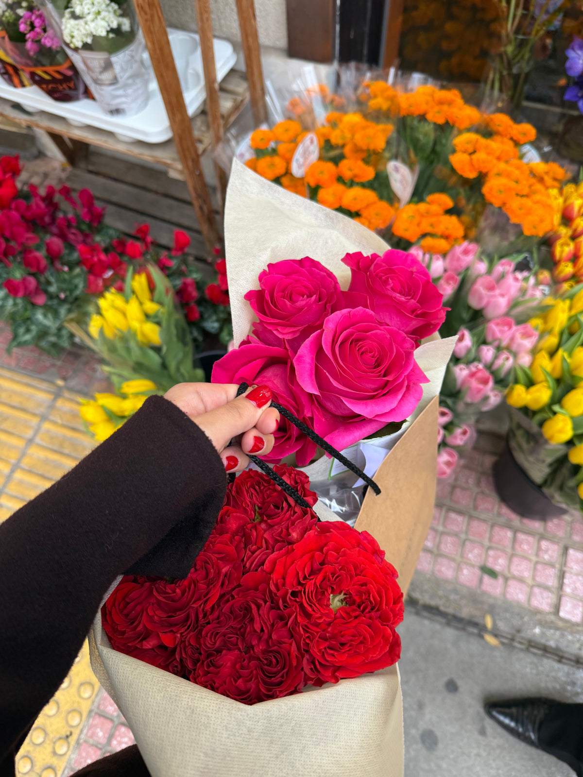 A hand holds a bouquet with bright pink and deep red roses at an outdoor flower market, surrounded by yellow, orange, and pink flowers.
