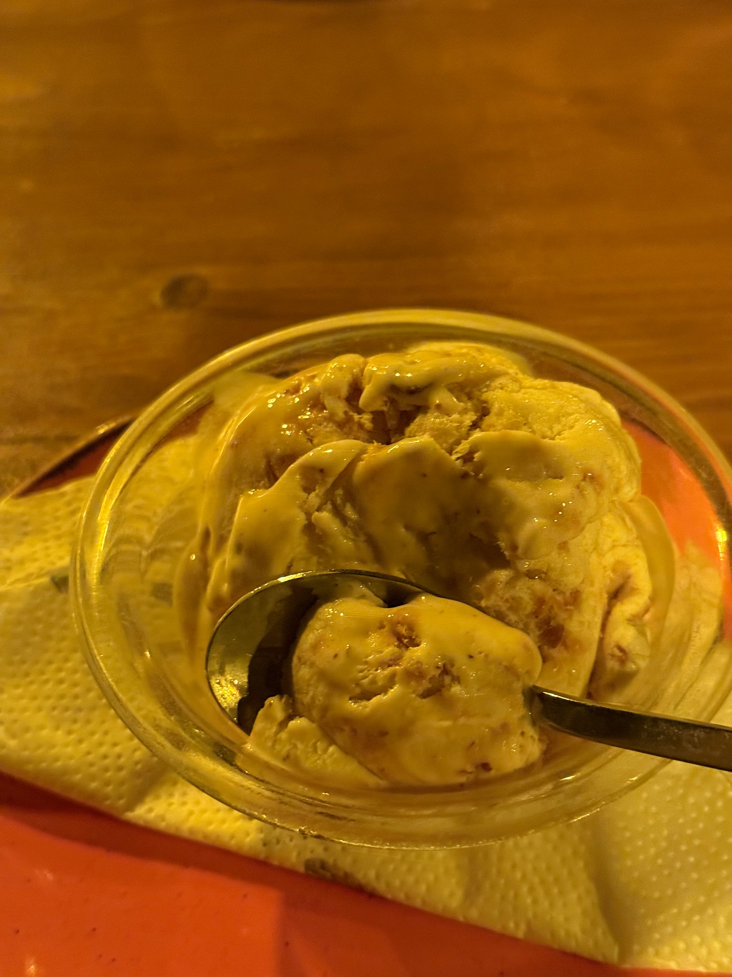 A bowl of creamy ice cream with visible chunks, served in a clear glass dish with a spoon resting inside, placed on a napkin over a red tray on a wooden table.