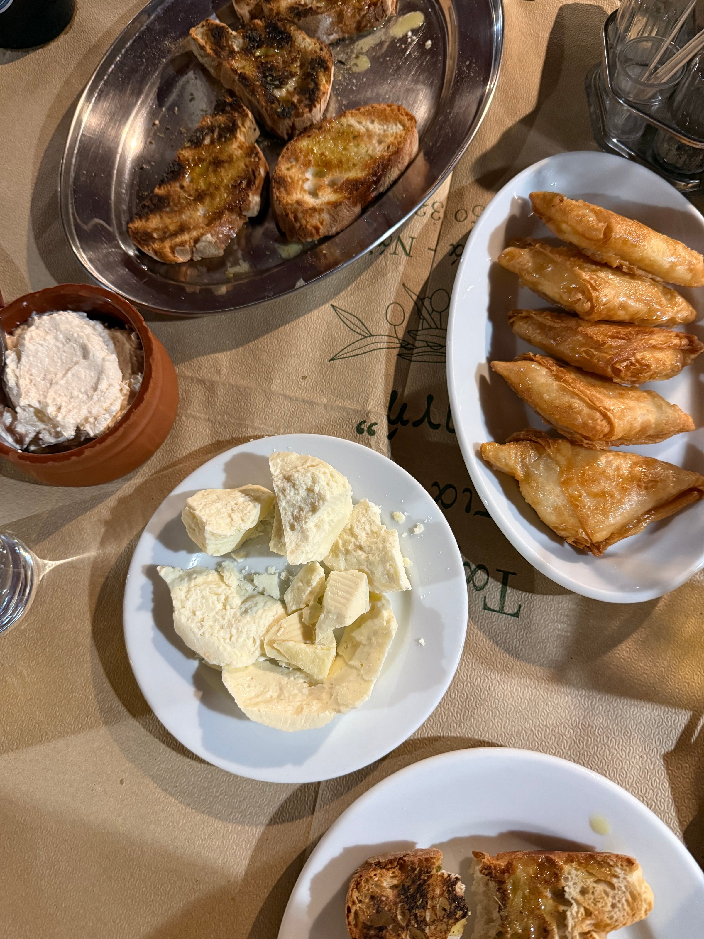 Plates of toasted bread, feta cheese, pastry triangles, and a small bowl of dip are arranged on a table covered with brown paper. A glass of water and utensils are also visible.